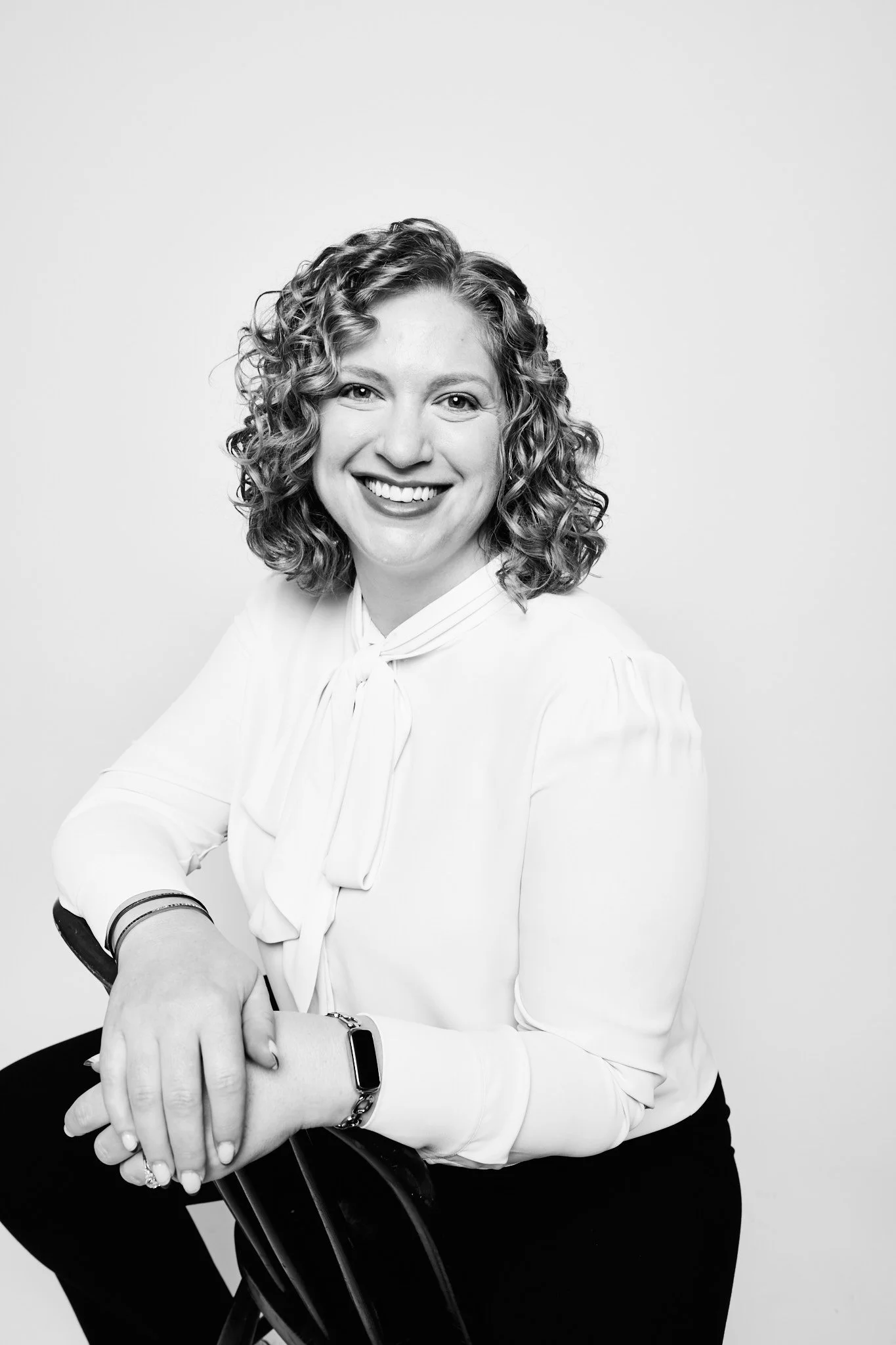 A woman with curly hair smiling, sitting with her arms crossed, wearing a white blouse and a smartwatch, in a black-and-white photo against a plain background.