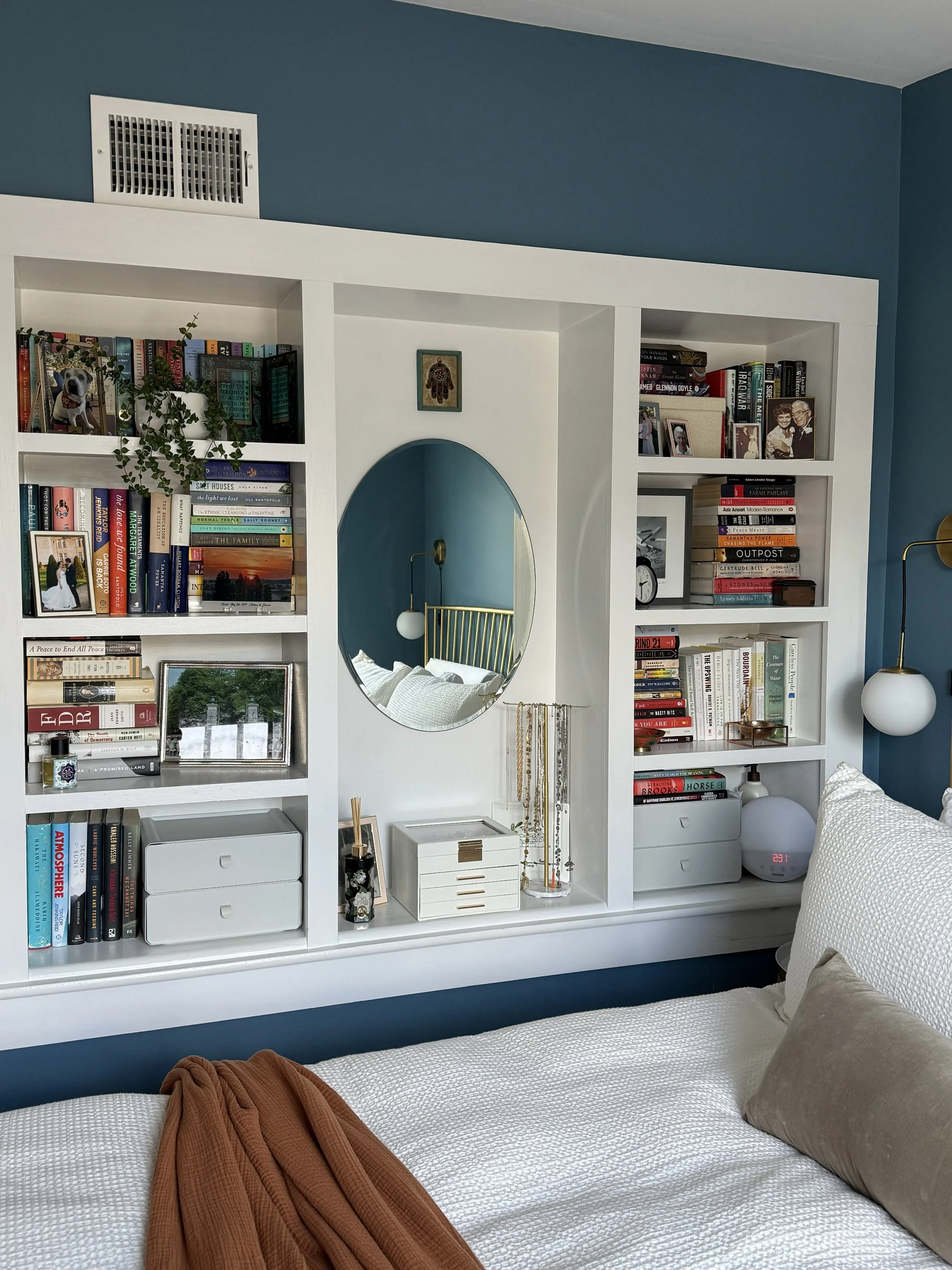 A white bed with a beige pillow and brown blanket in front of a blue wall with white built-in bookshelves, a mirror, and a small white dresser, decorated with books, photos, and decor items.