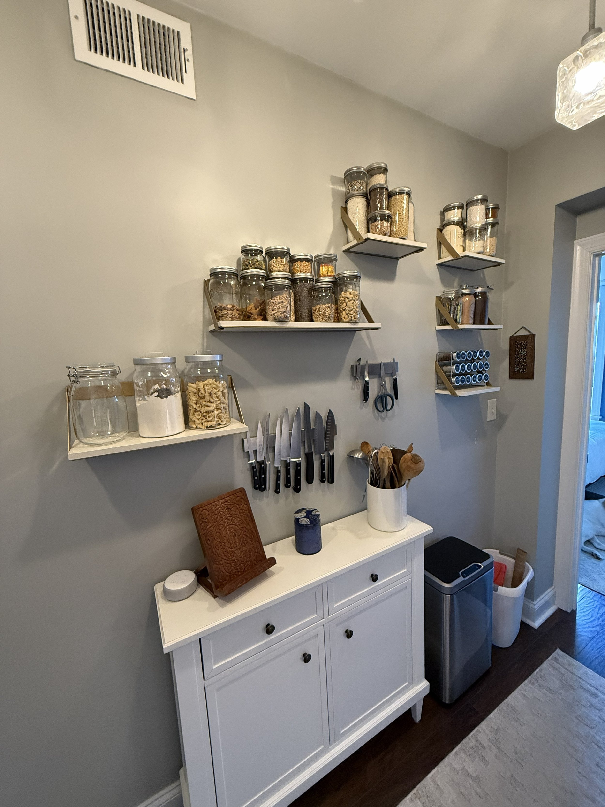A white sideboard with cabinets and drawers positioned against a light gray wall. Above it, there are three floating shelves mounted on the wall, each holding glass jars filled with various dry goods like grains, nuts, and spices. On top of the sideboard, there are large glass containers filled with pasta and other ingredients, a wooden recipe holder, and a small round device. To the right of the sideboard, there is a container holding utensils, a trash can, and a black step stool beside the wall. A doorway leads to another room with a glimpse of a bed.