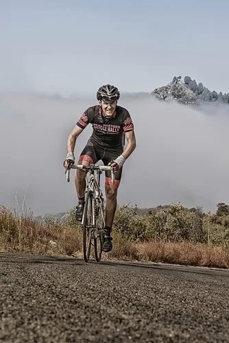 A cyclist in a black and red jersey riding a mountain bike on a dirt trail surrounded by fog and distant mountains.