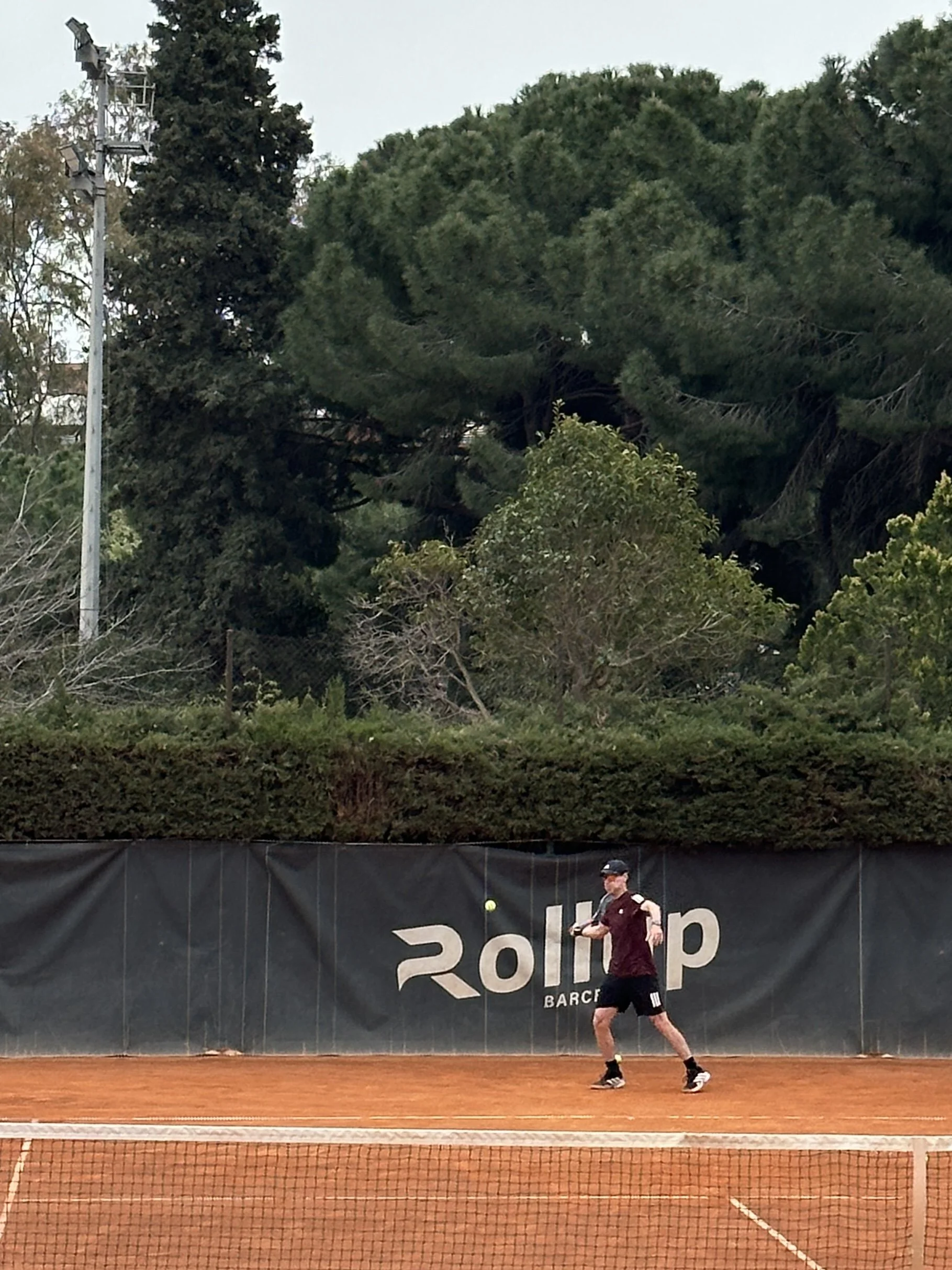 A person playing tennis on a clay court with a dark banner behind them and green trees in the background.