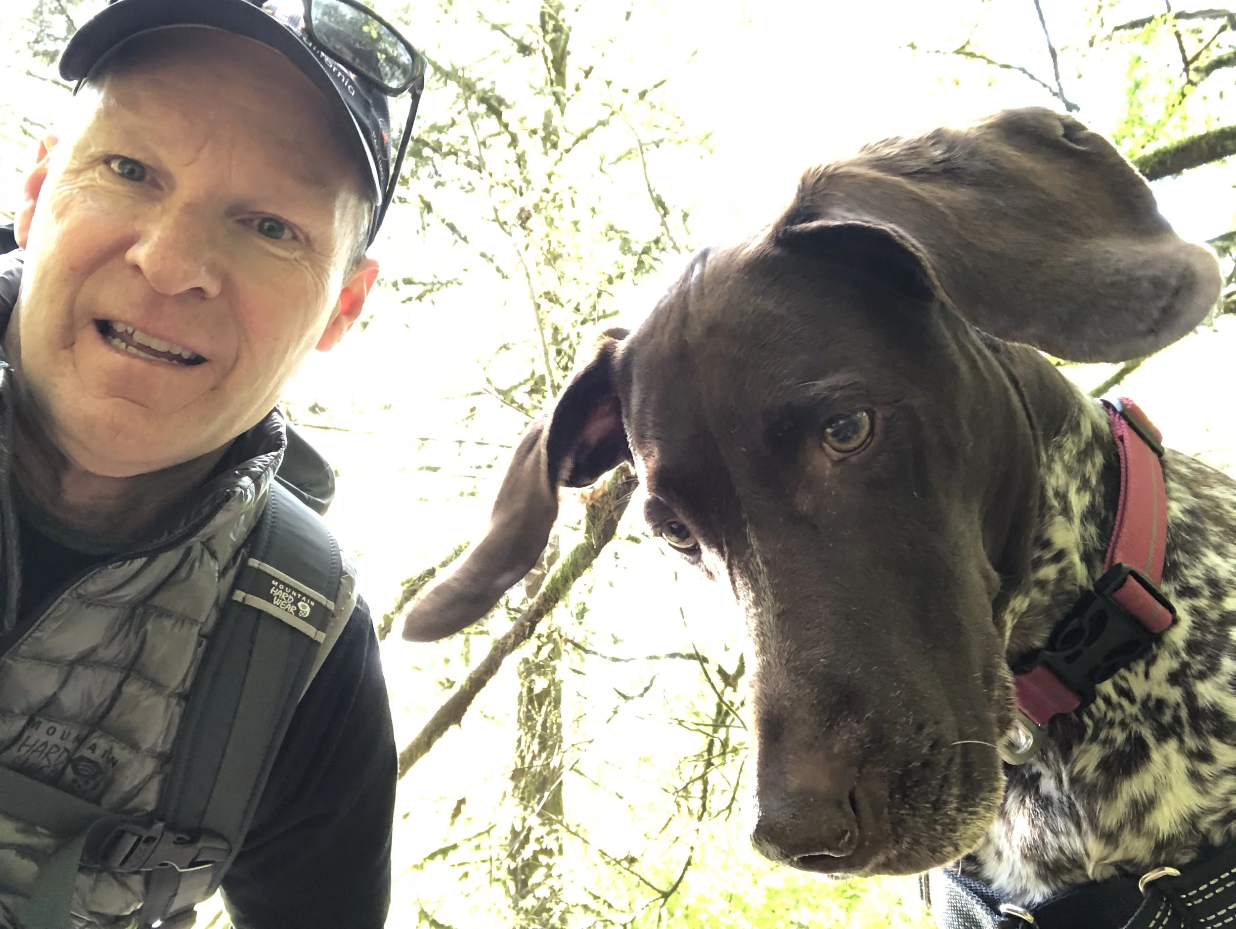 A man with a cap and outdoor jacket smiling next to a large dog with a brown and black coat, both outdoors under a tree.