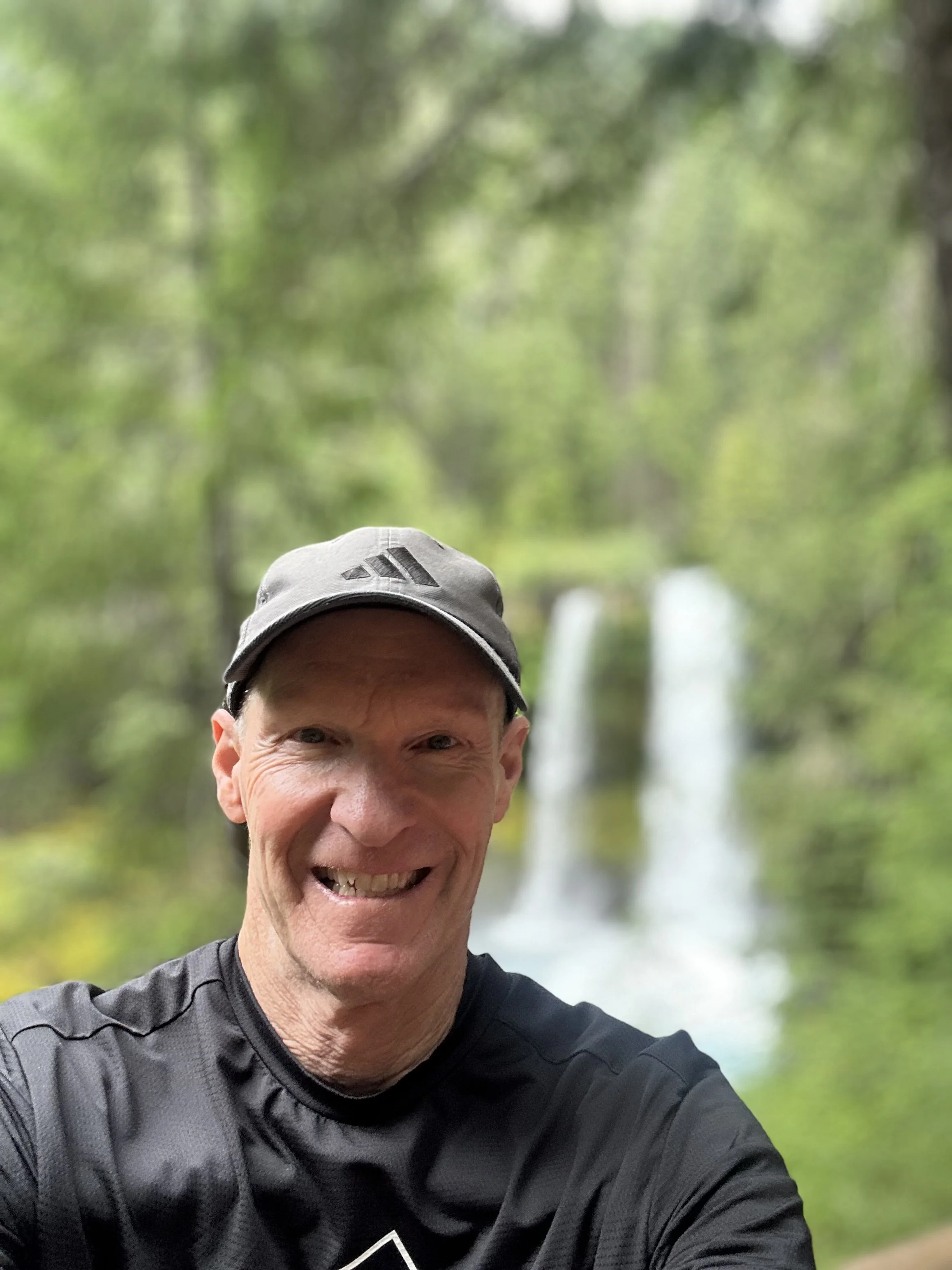 A man taking a selfie outdoors in a forested area with waterfalls in the background.