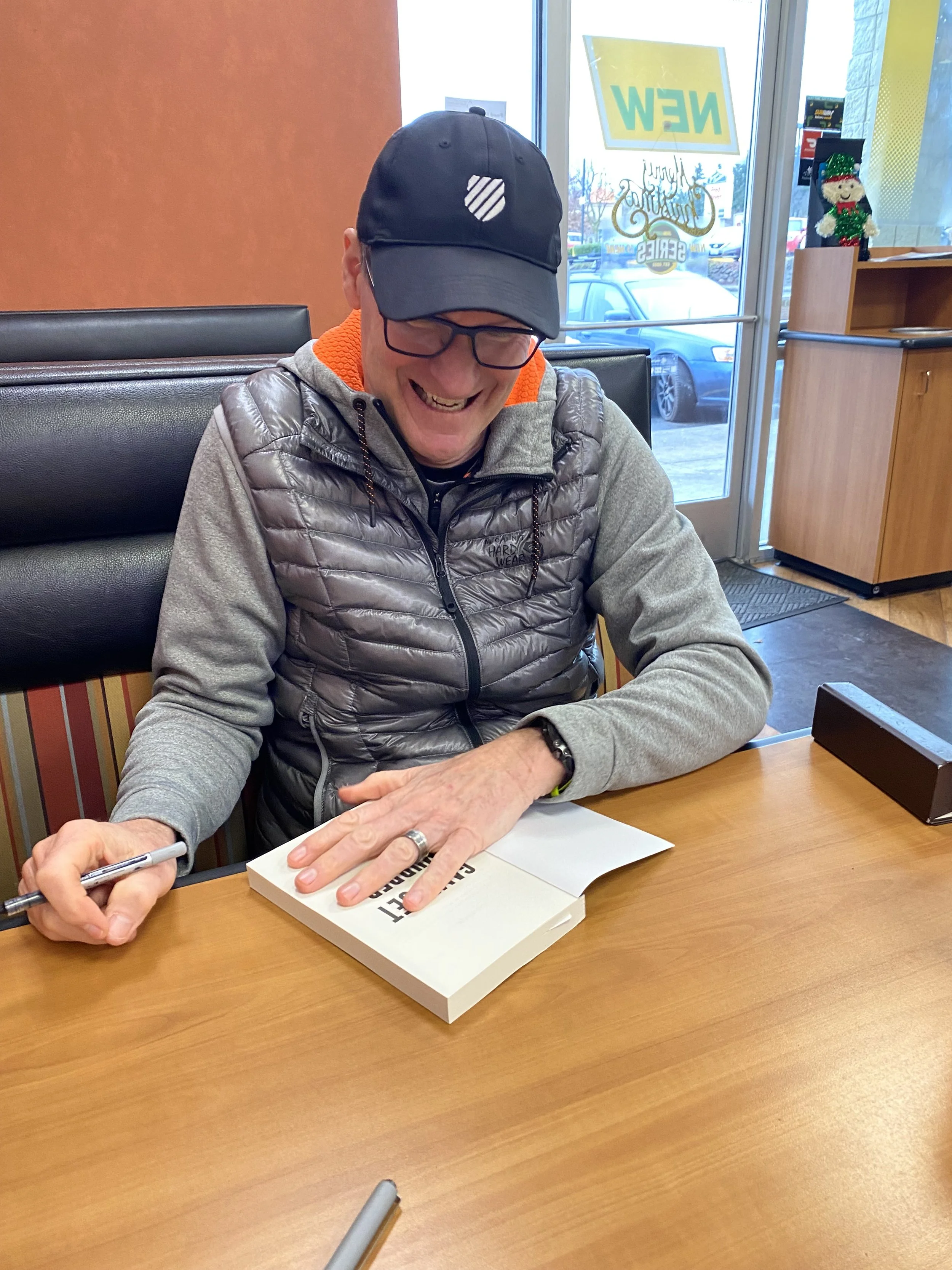 A man sitting at a table in a restaurant, smiling and signing a book with a pen. He is wearing glasses, a black cap, a gray puffer vest, and a gray hoodie with orange lining. There is a small Christmas decoration on a shelf near the window, and a sign in the window saying 'NEW'.