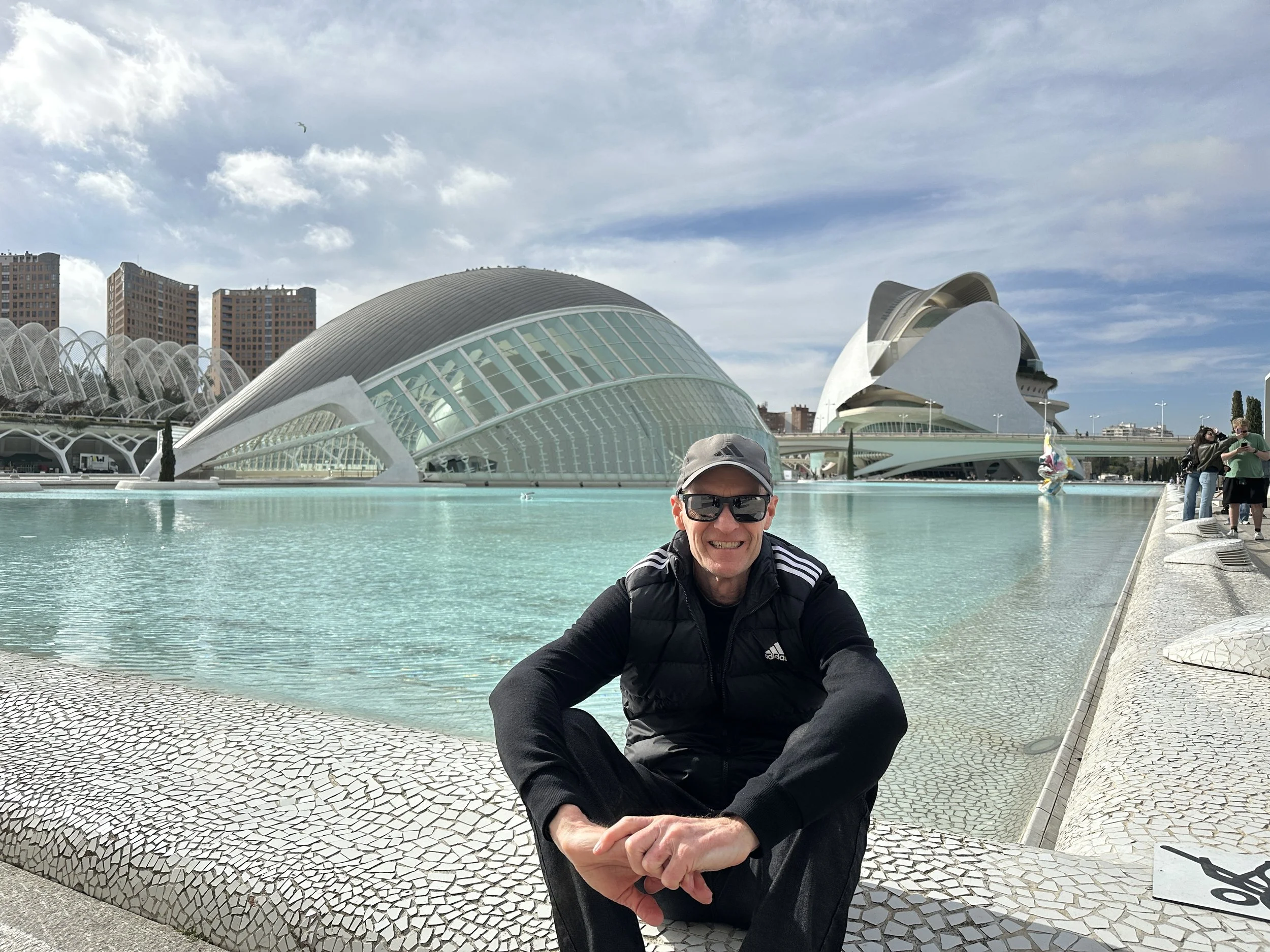 A man smiling, wearing sunglasses, a black jacket, and a light-colored cap, sitting near a pool with modern white buildings and cityscape in the background.