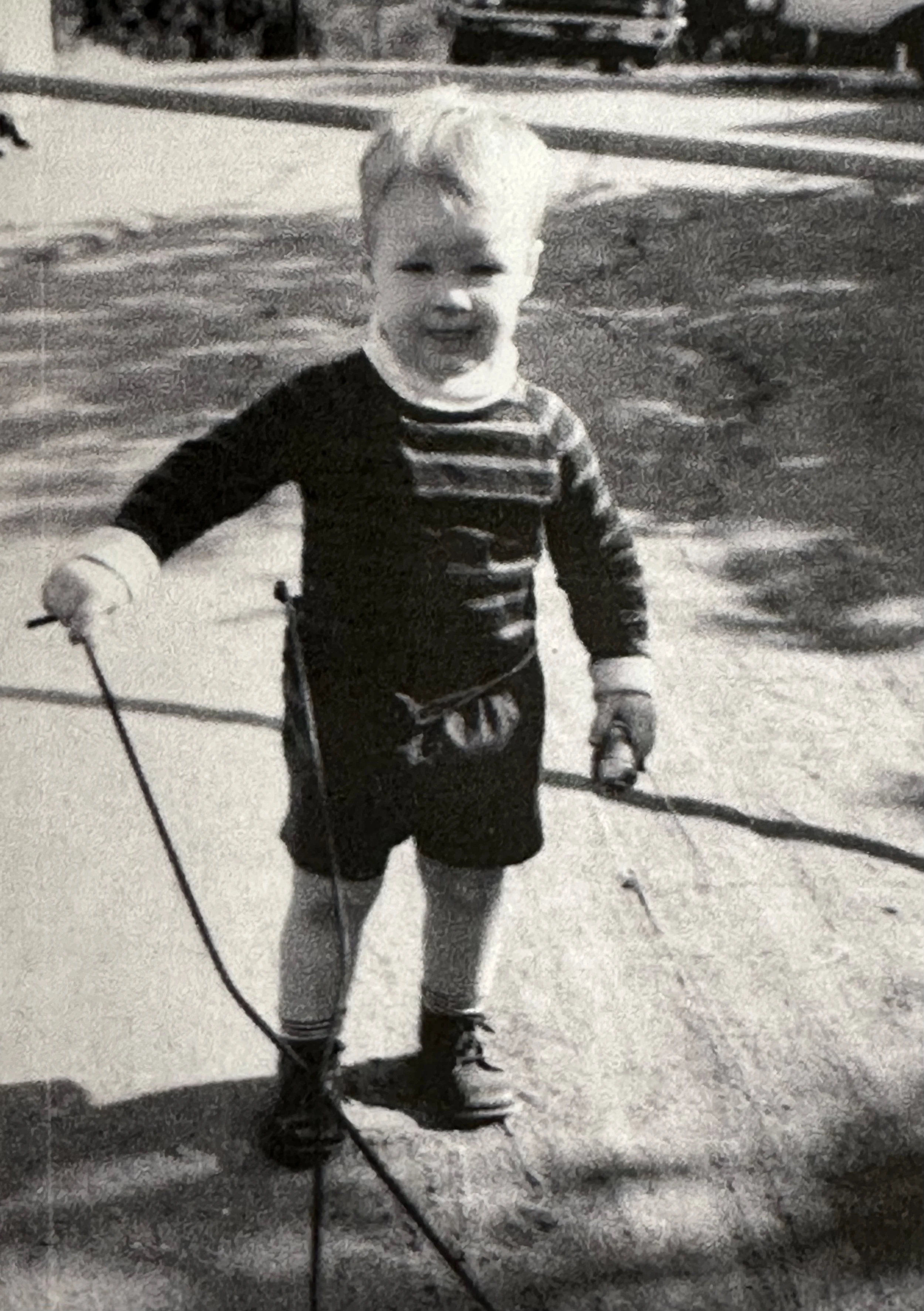 A young boy standing outdoors, holding two small dumbbells, with a water body and a bridge or structure in the background, in black and white.