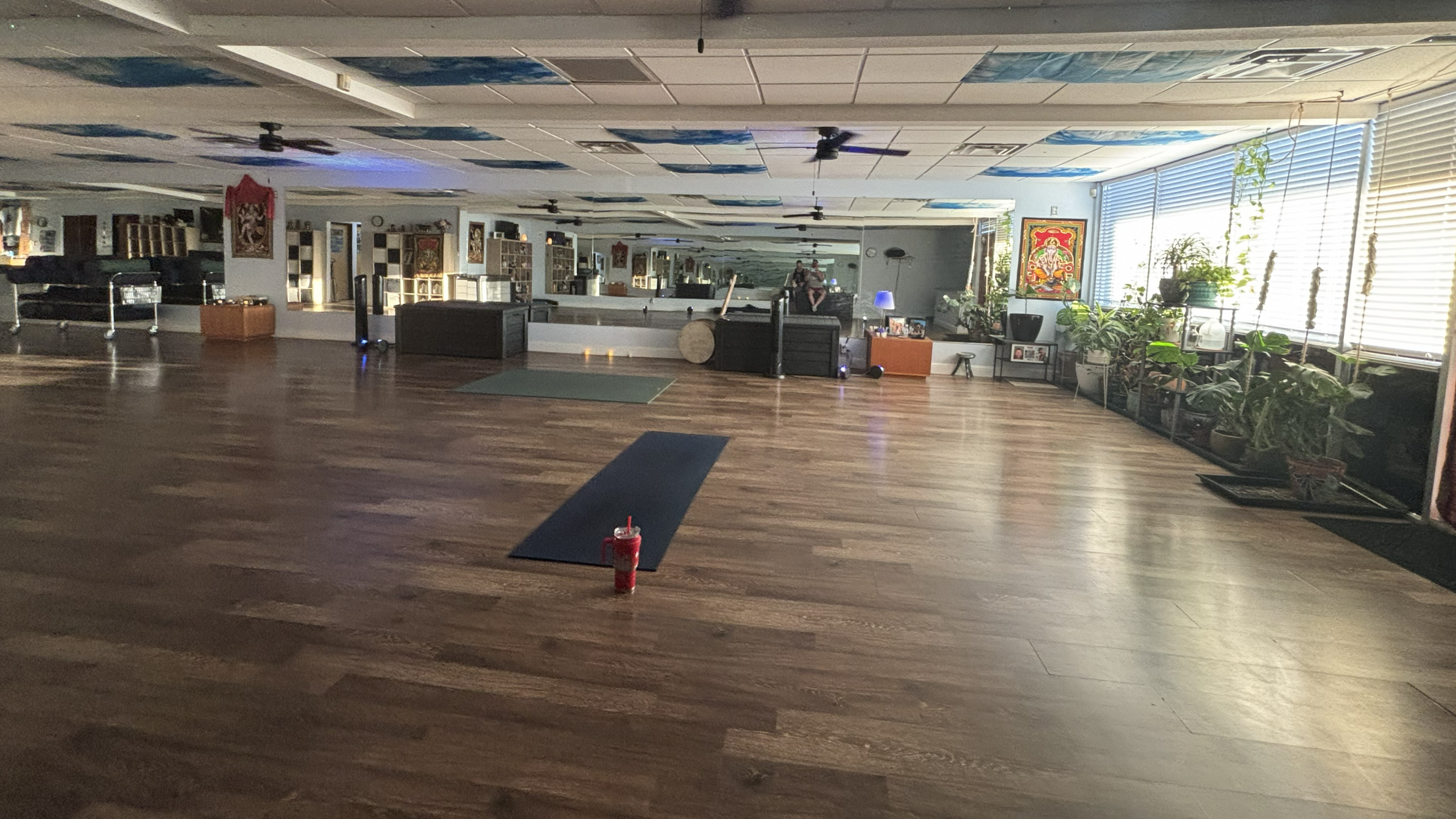 A large indoor space with hardwood floors, decorated with plants and artwork, and a large mirror on one wall reflecting the room. A yoga mat and a red cup are on the floor, and there are some chairs and shelves along the walls.