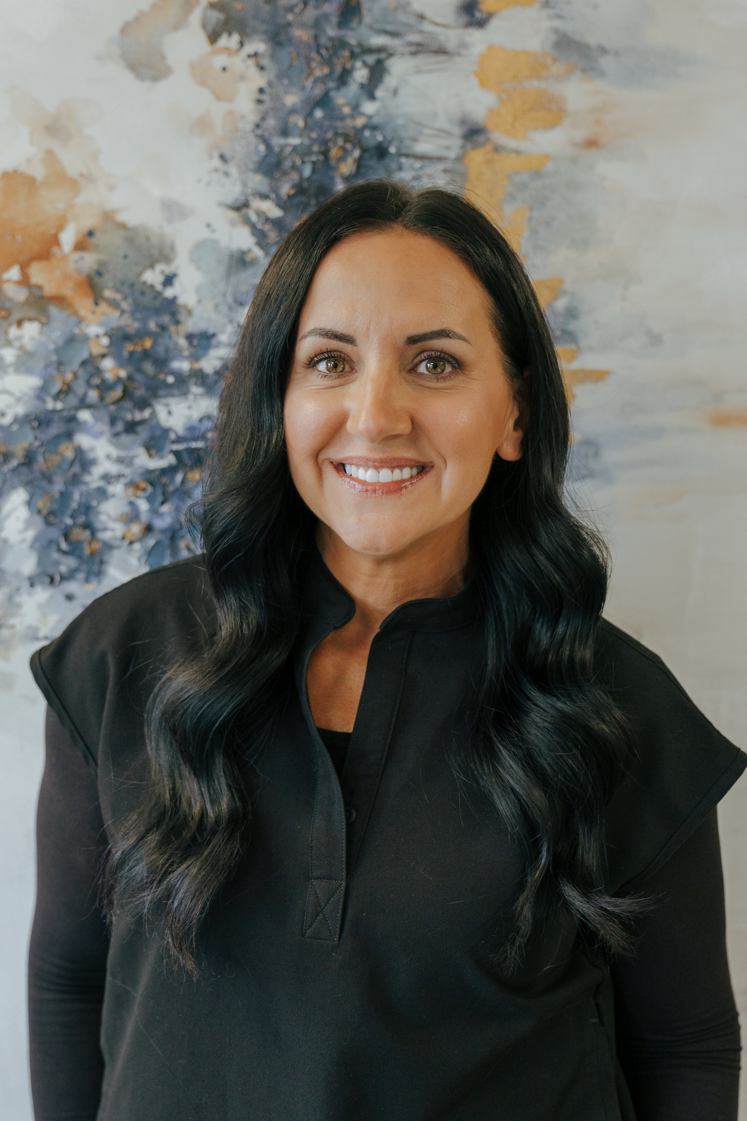 A woman with dark brown hair, wearing a black top, smiling indoors.