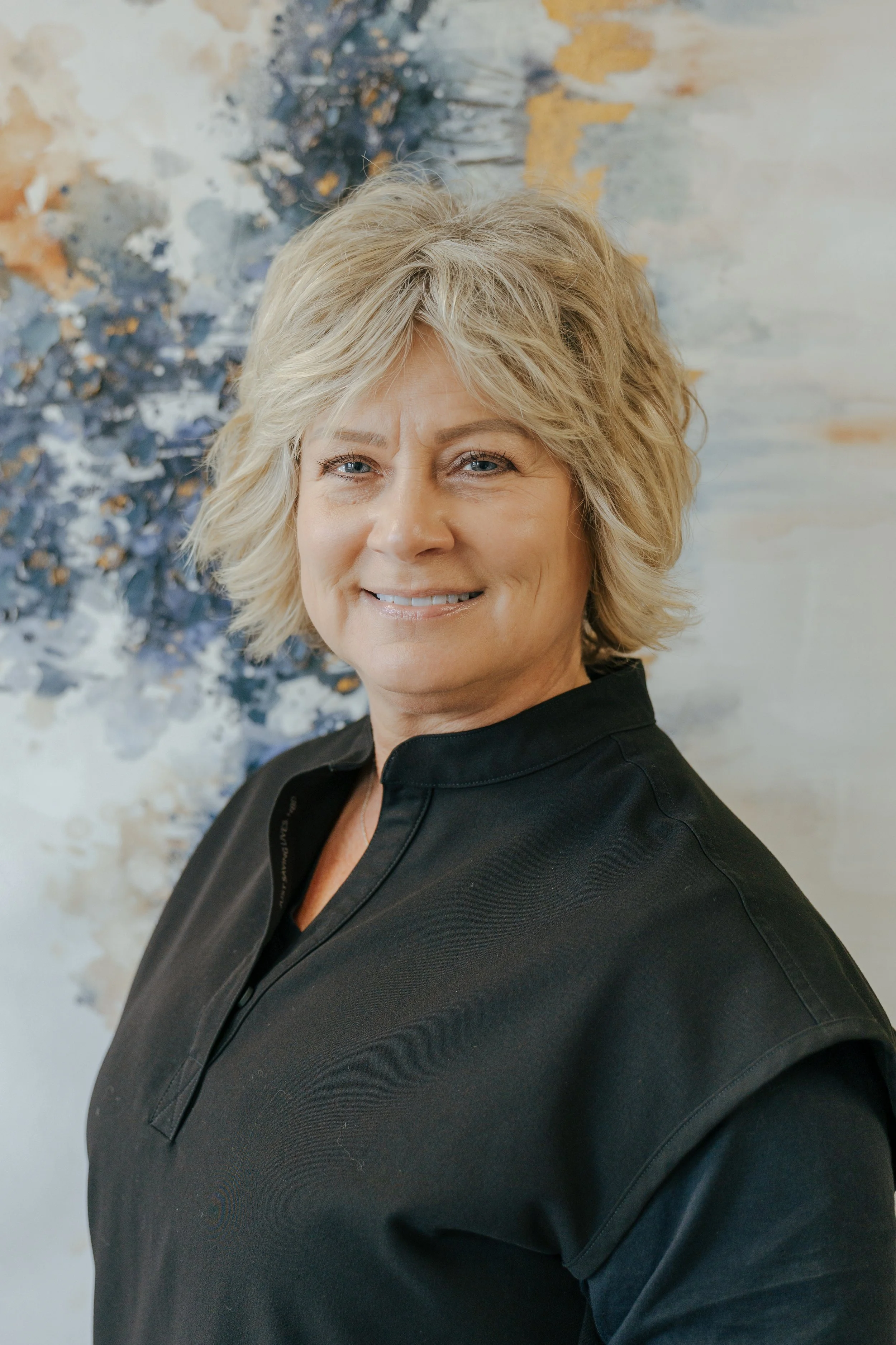 A smiling woman with short blonde hair, wearing a dark sweater and a necklace, standing indoors near a window with a scenic landscape of green fields and trees outside under a partly cloudy sky.