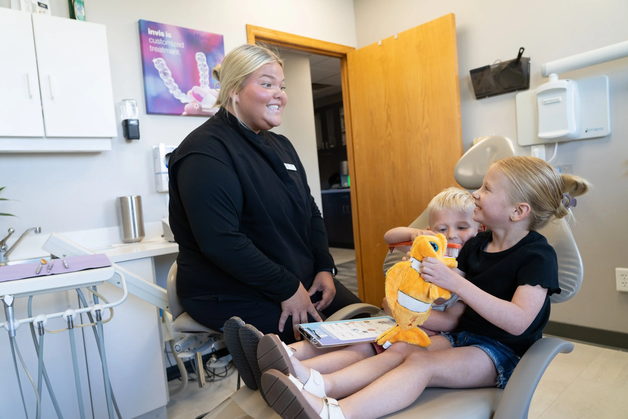 A young girl in a dental chair holding a tiger plush toy, smiling at a female dental professional who is sitting nearby and smiling back, with two young children in the background in a dental office.