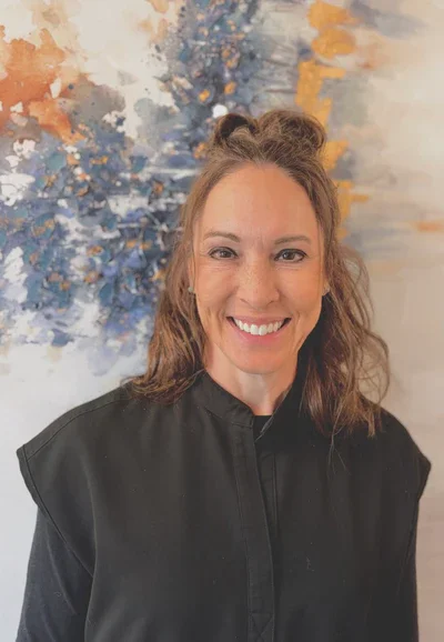 A woman with light skin and brown hair styled in a half-up bun, smiling, wearing a black top, standing in front of an abstract colorful painting with shades of blue, white, orange, and yellow.