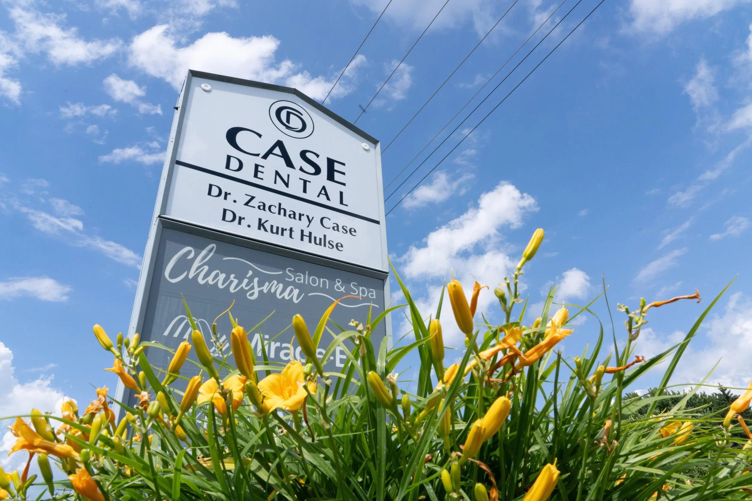 A tall sign for a dental practice with the names Dr. Zachary Case and Dr. Kurt Hulse, set against a bright blue sky with scattered clouds, surrounded by yellow daylilies in full bloom.