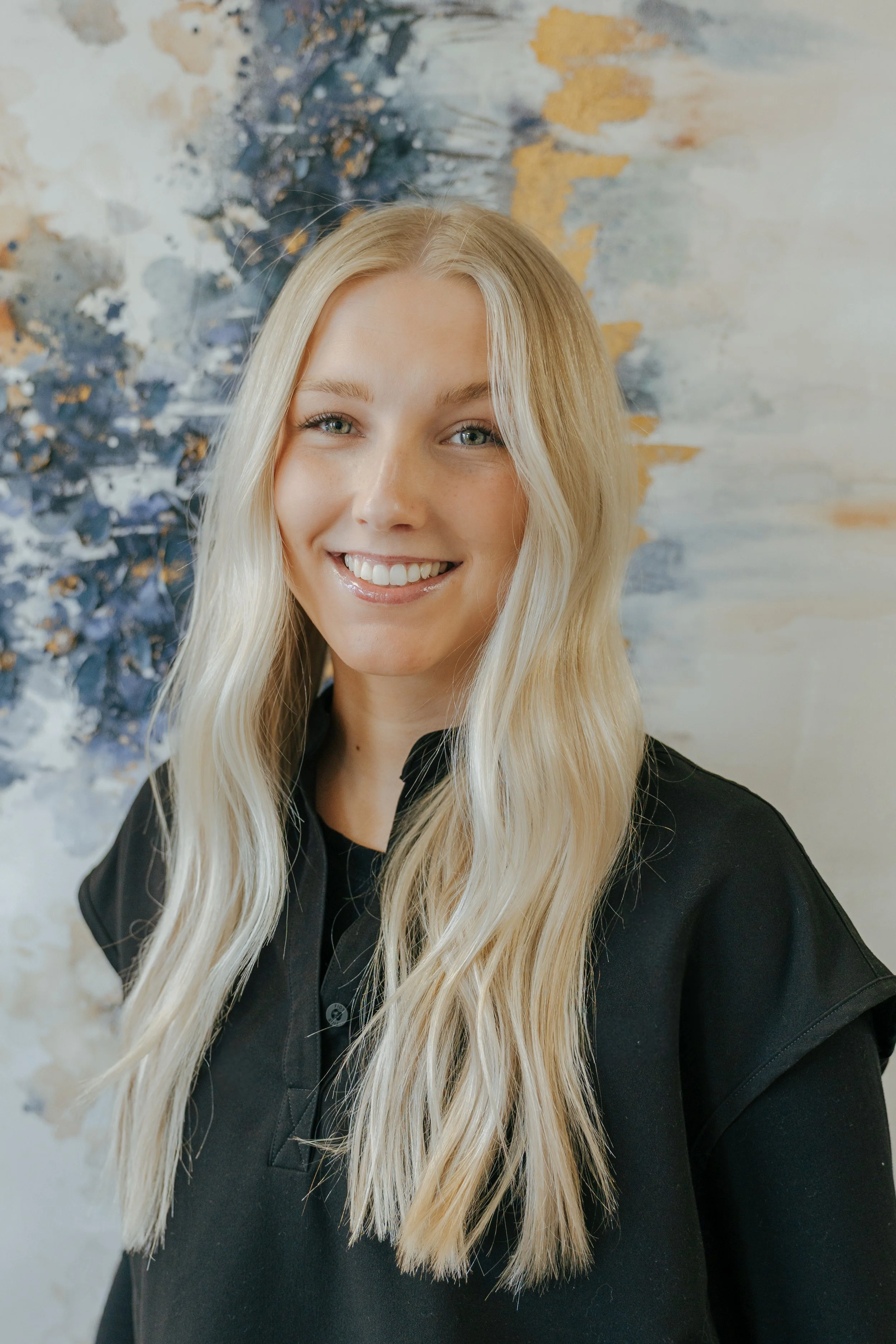 Young woman with long blonde hair and a black zip-up jacket smiling in front of an abstract painting with blue, white, and gold textures.
