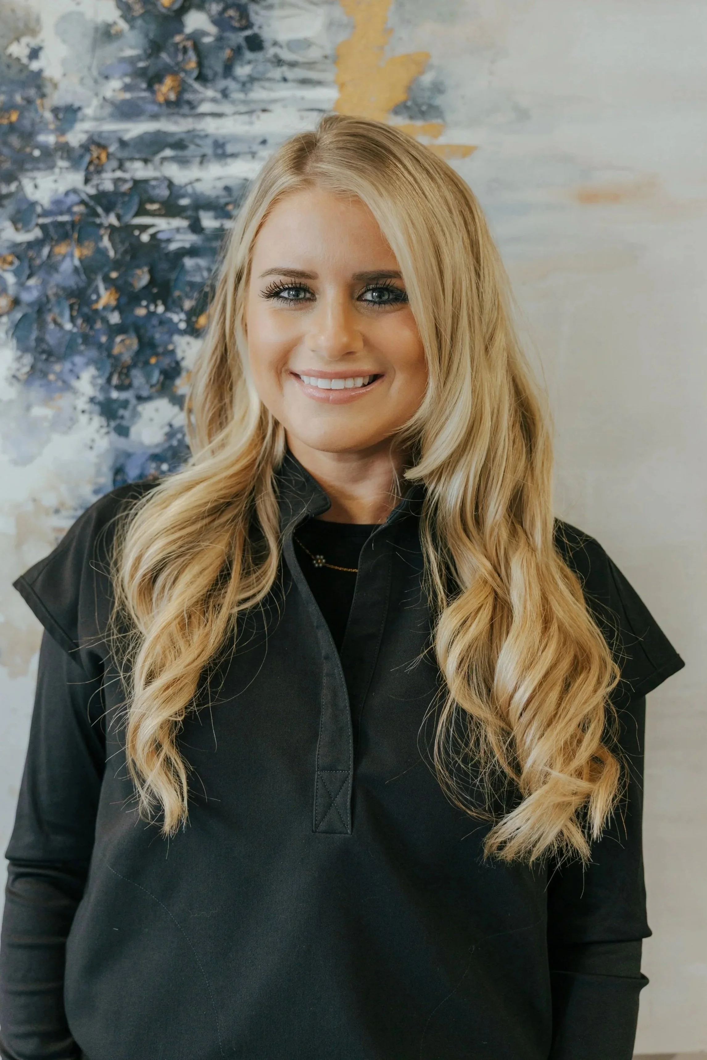 Young woman with long blonde hair, smiling, standing in front of a colorful abstract floral painting.