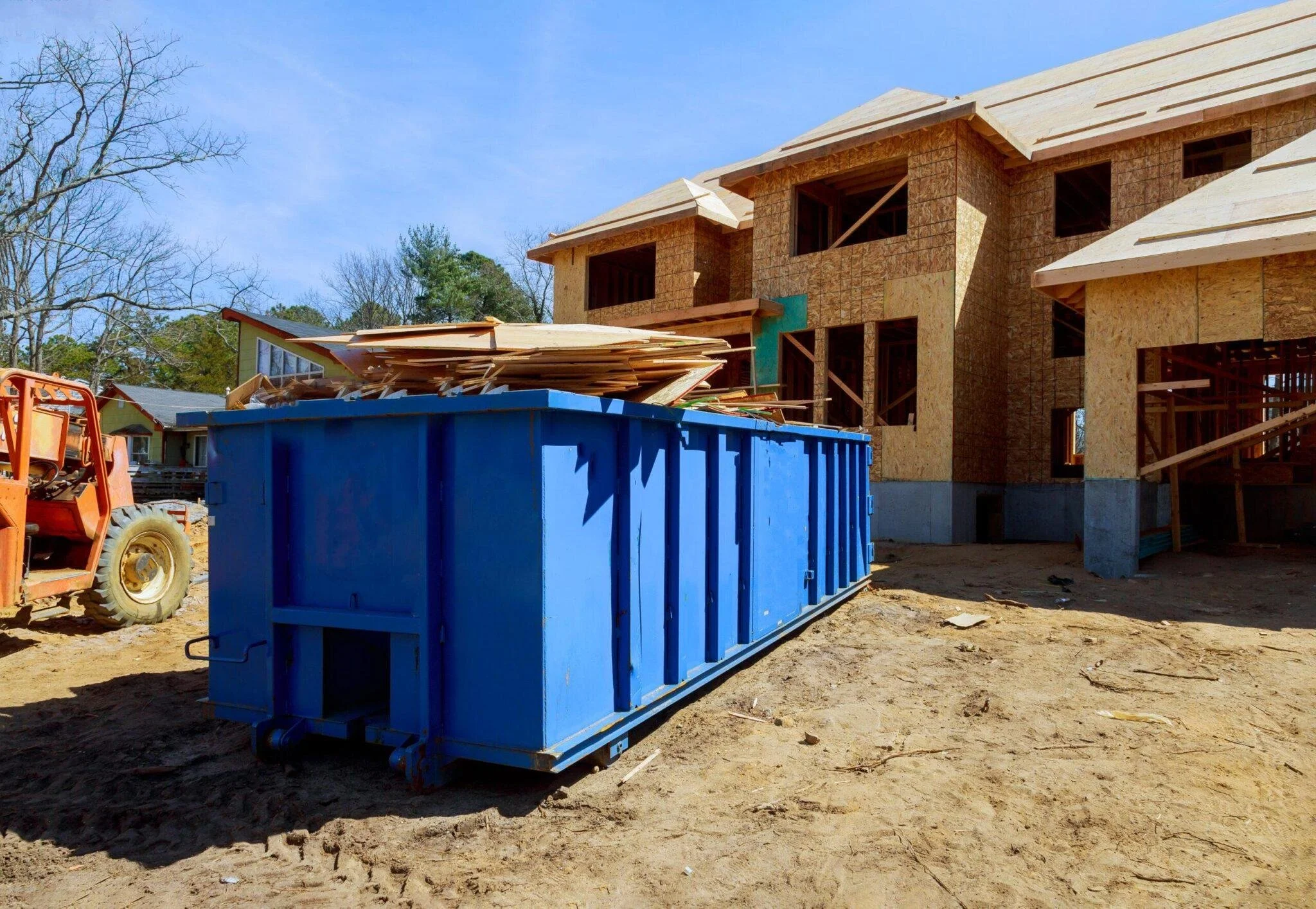 Construction site with a partially built two-story house surrounded by trees, a blue dumpster filled with wooden debris, and construction equipment on dirt ground.