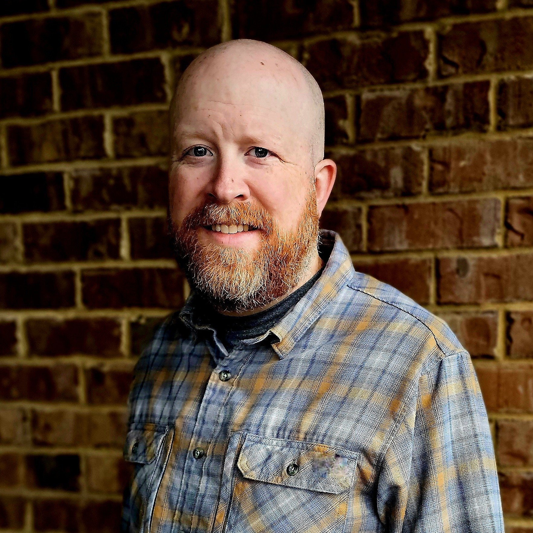 A man with a reddish beard and short hair smiling, wearing a plaid shirt, standing in front of a brick wall.