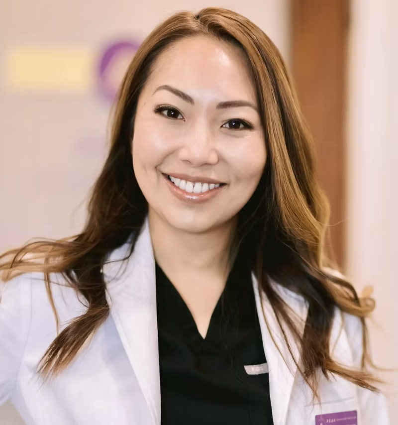A woman with long, wavy brown hair and a bright smile, wearing a white medical coat over black scrubs, standing indoors with a blurred background.