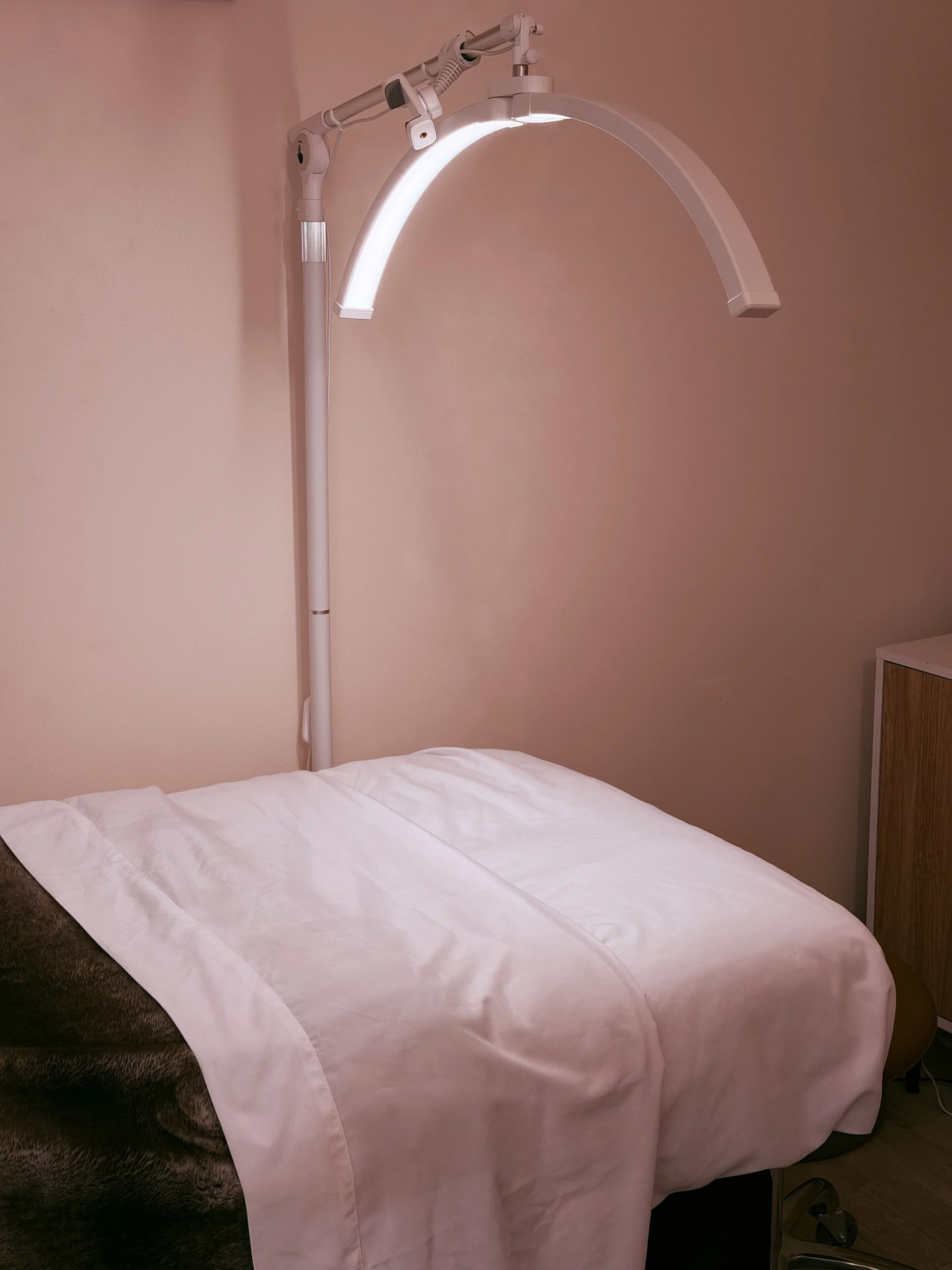 A hospital room bed with white bed linens and a padded headboard, adjacent to a medical examination lamp attached to the wall.