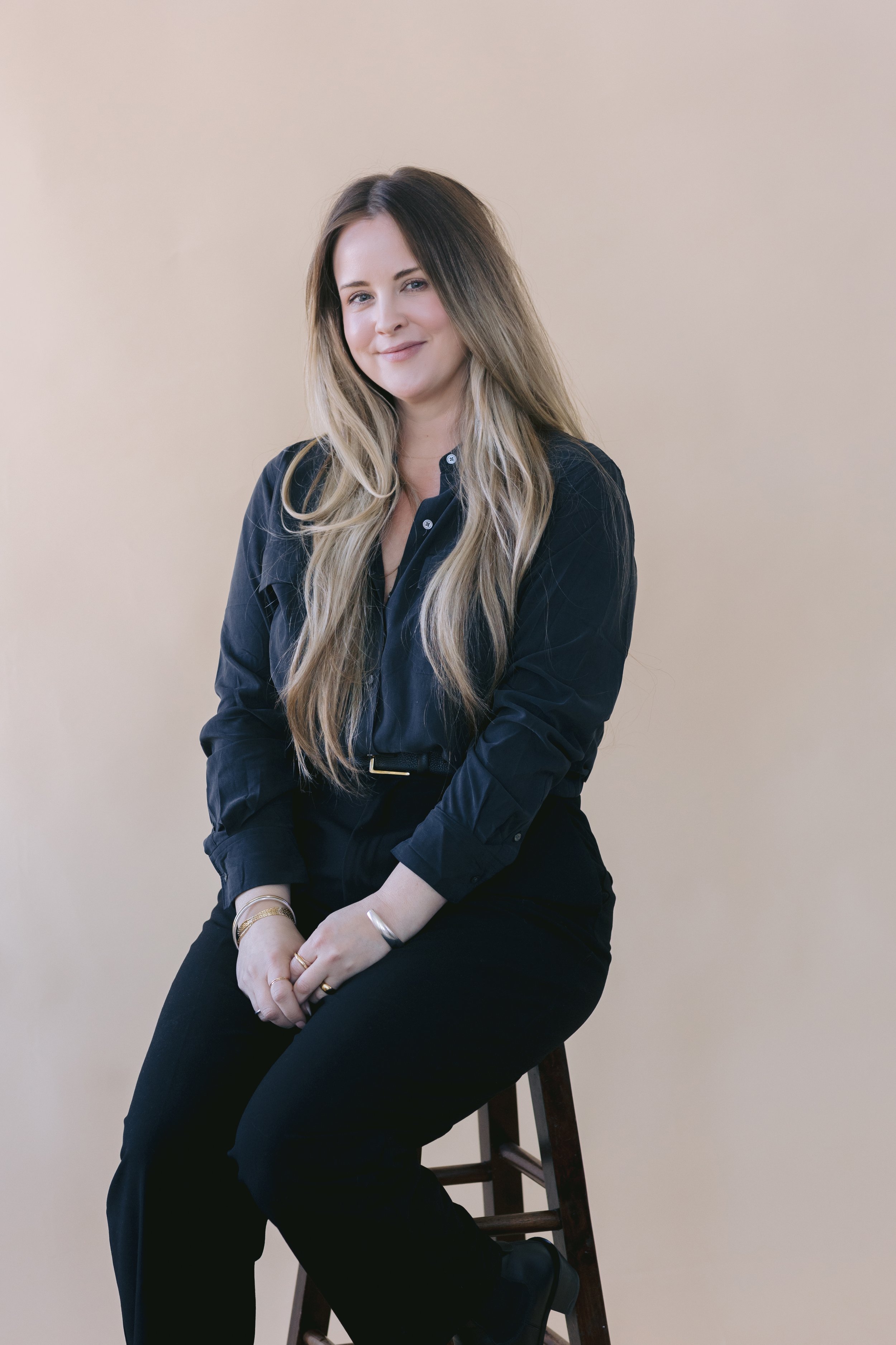 A woman with long, blond hair sitting on a wooden stool, wearing a black shirt and black pants against a plain beige background.