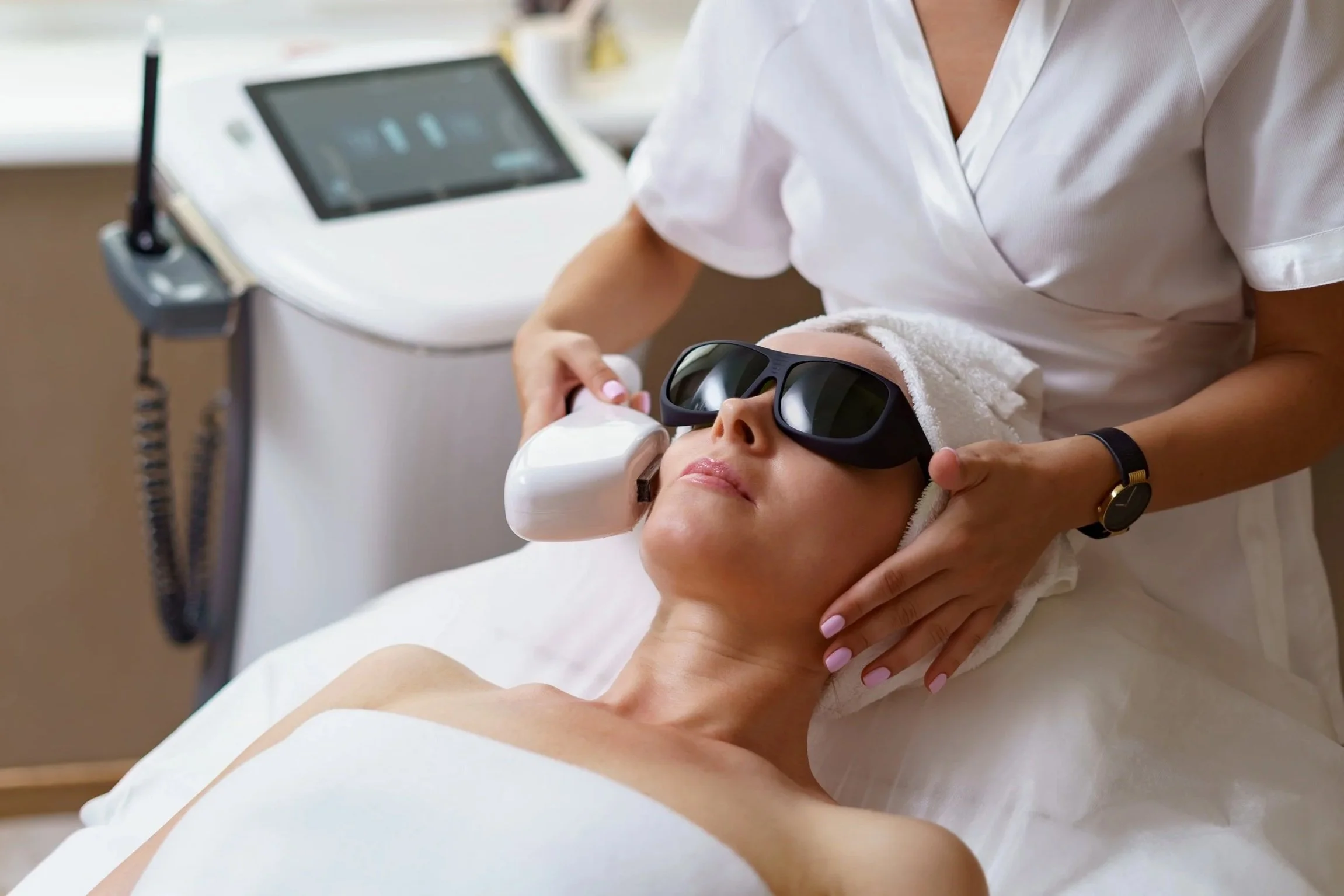 A woman receiving a facial treatment with protective glasses, lying on a bed covered with a white towel, while an aesthetician holds a device near her face in a spa or clinic setting.