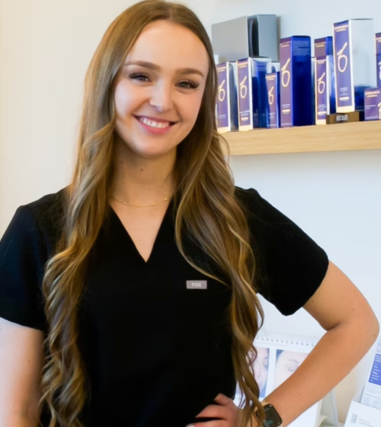 A smiling woman with long wavy hair wearing a black uniform with a name tag that says 'FIGS' in a professional setting, standing in front of a shelf with boxes of products.