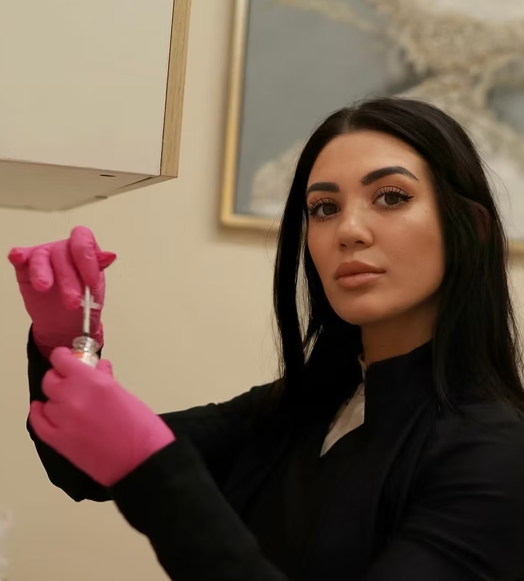 A woman with black hair and a serious expression, wearing pink gloves, is preparing a syringe with a small vial in an indoor setting.