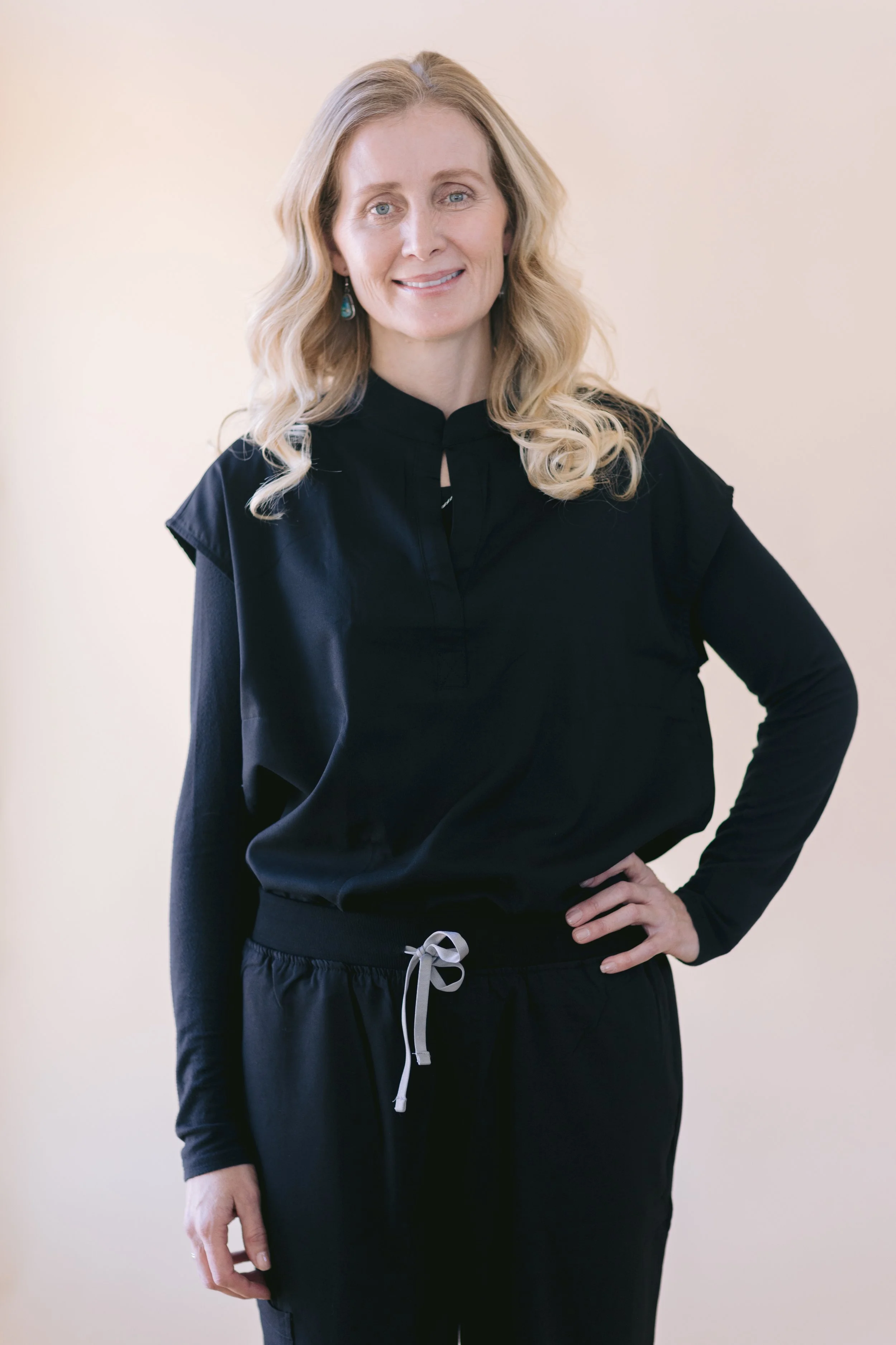 A woman with blonde wavy hair wearing a black long sleeve top and black drawstring pants, standing with one hand on her hip, smiling, against a plain light-colored background.