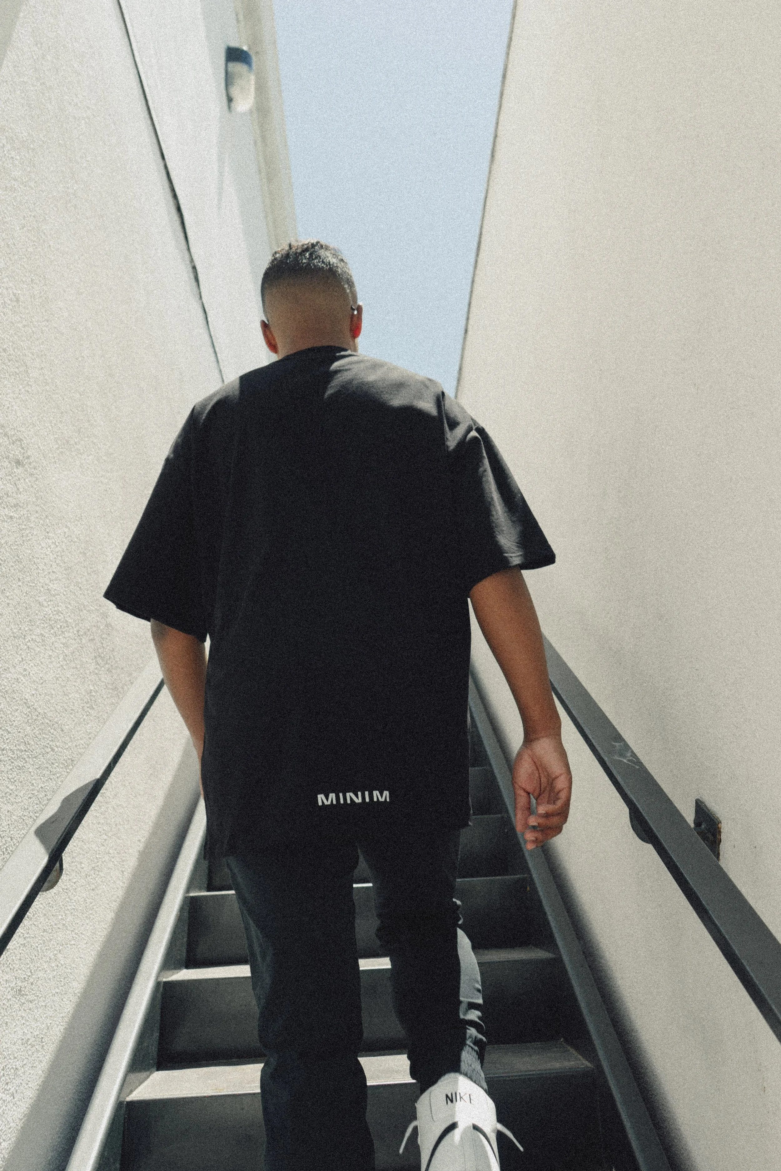 A person walking up a staircase in an airport or similar setting, viewed from behind. The person is wearing a black shirt, black pants, and white Nike sneakers with the name 'NIKI' visible on the shoes. The staircase is enclosed by cream-colored walls and is lit by natural light from above.