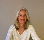 A smiling woman with shoulder-length blonde hair wearing a white top, sitting in front of a plain gray wall.