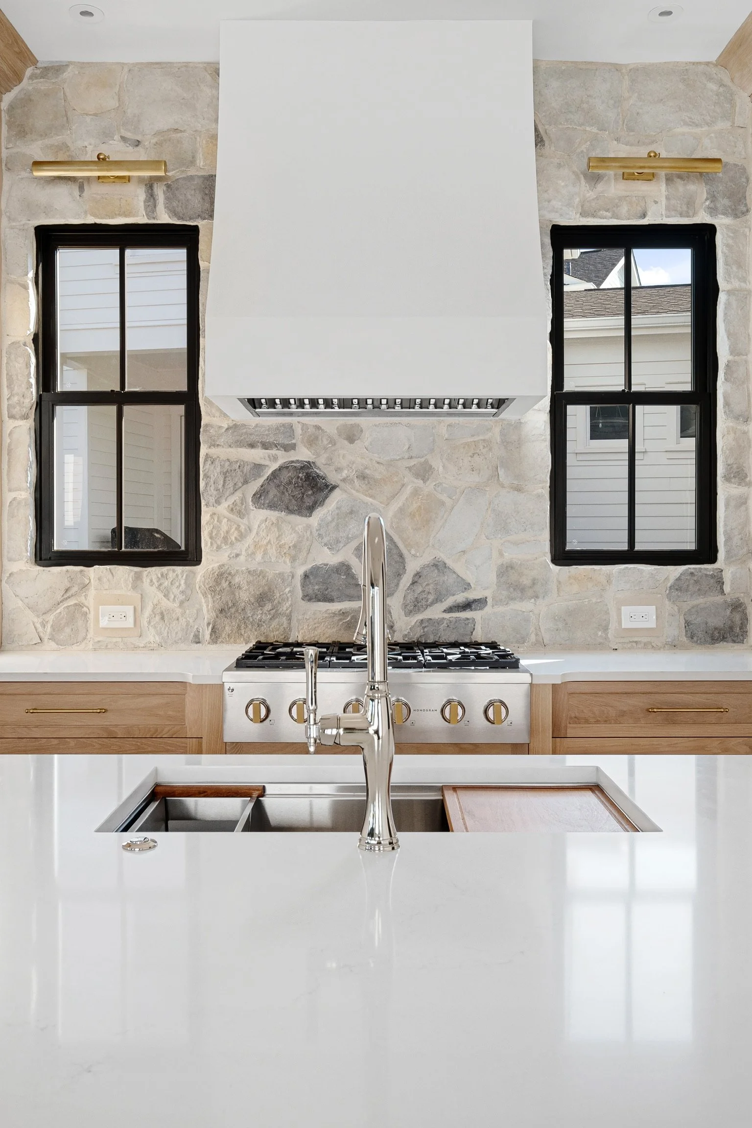 Modern kitchen with a white island counter, a stainless steel sink and faucet, wooden cabinets, a stone accent wall, and two black-framed windows with a view of the house exterior. DIY, DESIGN, STONE, KITCHEN STONE, HOOD VENT, PLASTER HOOD VENT