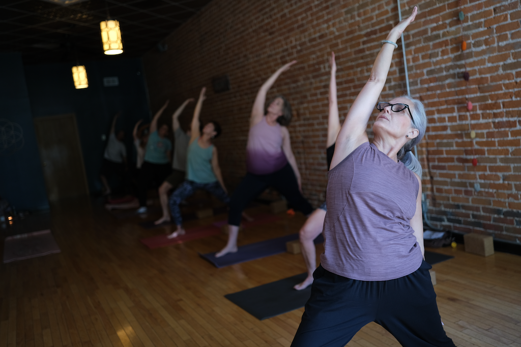 Older woman practicing yoga with stretch pose in a brick-walled studio, with other women behind her also doing yoga stretches.