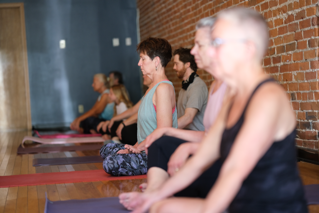 People participating in a yoga or meditation class in a studio with a brick wall, sitting cross-legged with eyes closed and hands resting on knees.