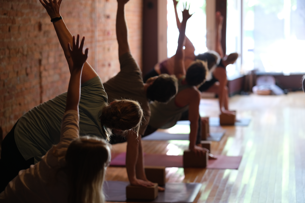 Multiple people practicing yoga in a studio, in side plank pose with raised arms, on yoga mats with blocks, against a brick wall and bright natural light.