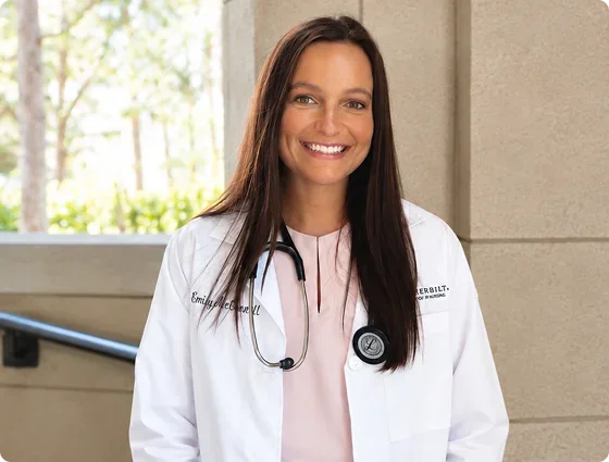 A smiling female doctor or medical professional with long brown hair, wearing a white lab coat and stethoscope, standing indoors near a window with trees outside.