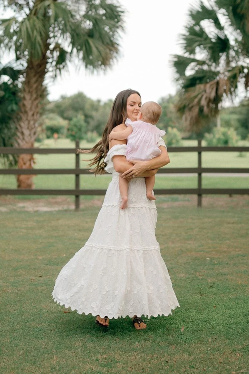 A woman holding a baby girl outdoors on a green lawn with palm trees in the background.