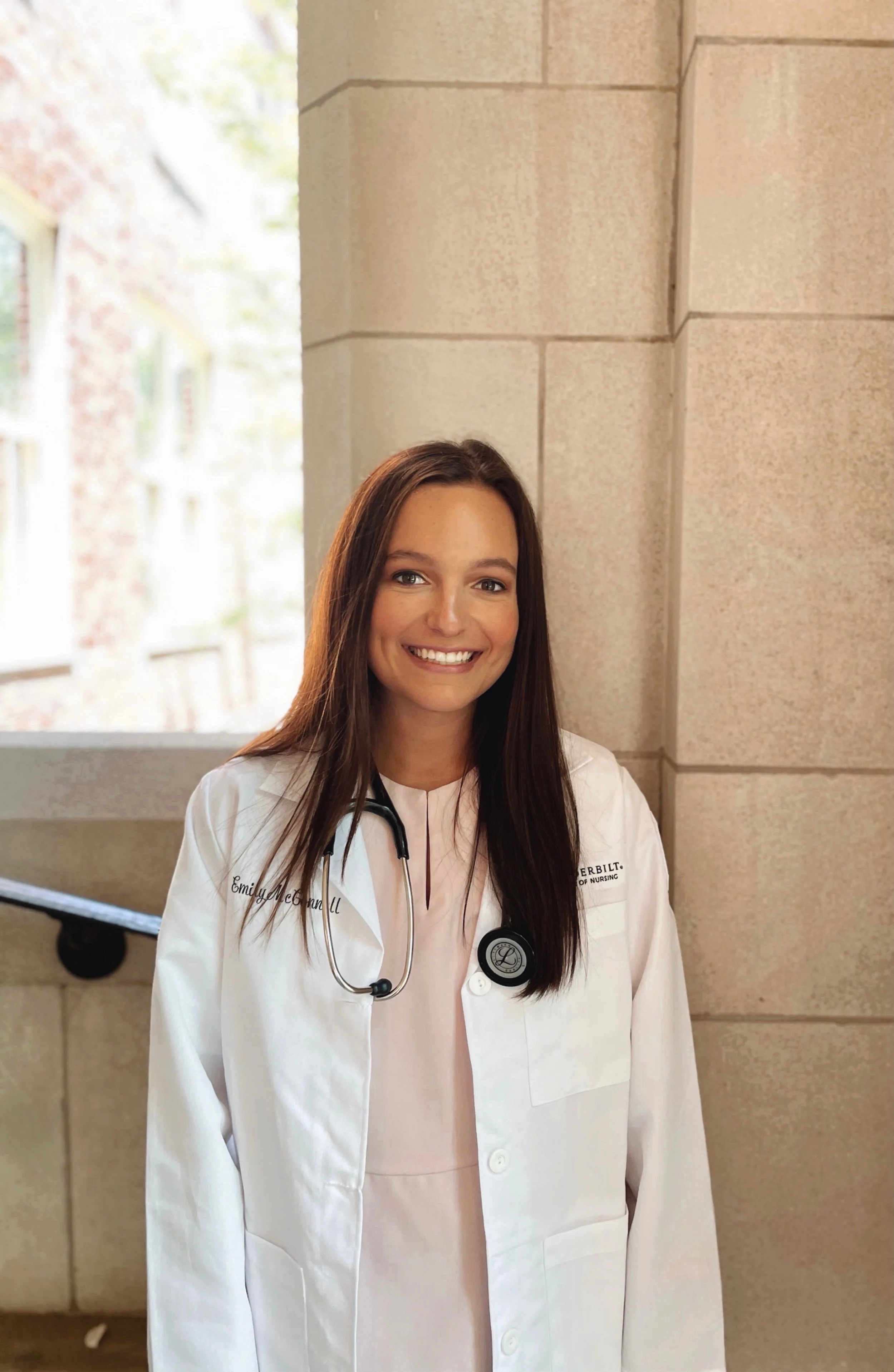 A young woman with long brown hair, wearing a white medical coat with a stethoscope, standing indoors against a beige stone wall with a window to her left.