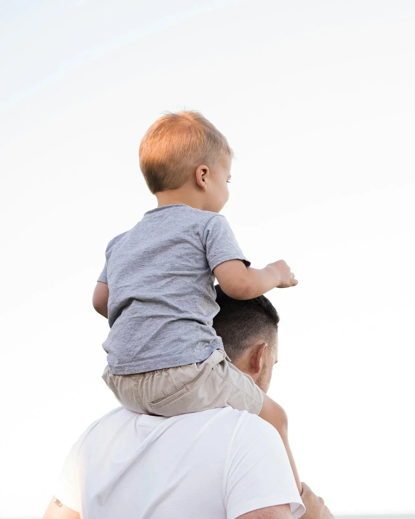 A young boy with ginger hair riding on the shoulders of a man with short dark hair and a beard, against a plain white background.
