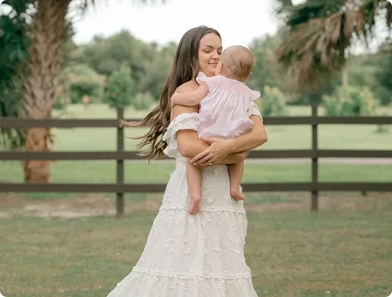 A woman holding a young child outdoors in a grassy area with trees and a wooden fence in the background.