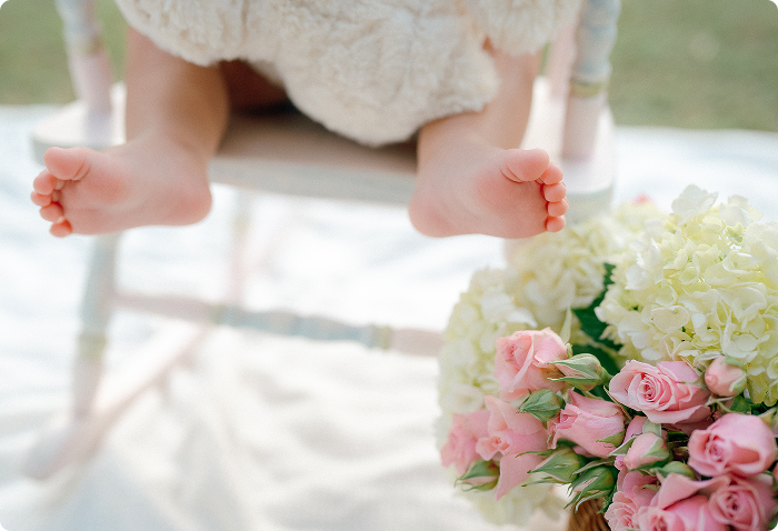 Close-up of a baby's bare feet hanging off the edge of a white wooden chair, with a bouquet of pink roses and white flowers on the floor nearby.