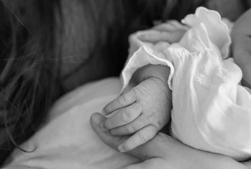 Close-up of an adult holding a baby's hand, with the baby's hand resting on the adult's arm, in black and white.