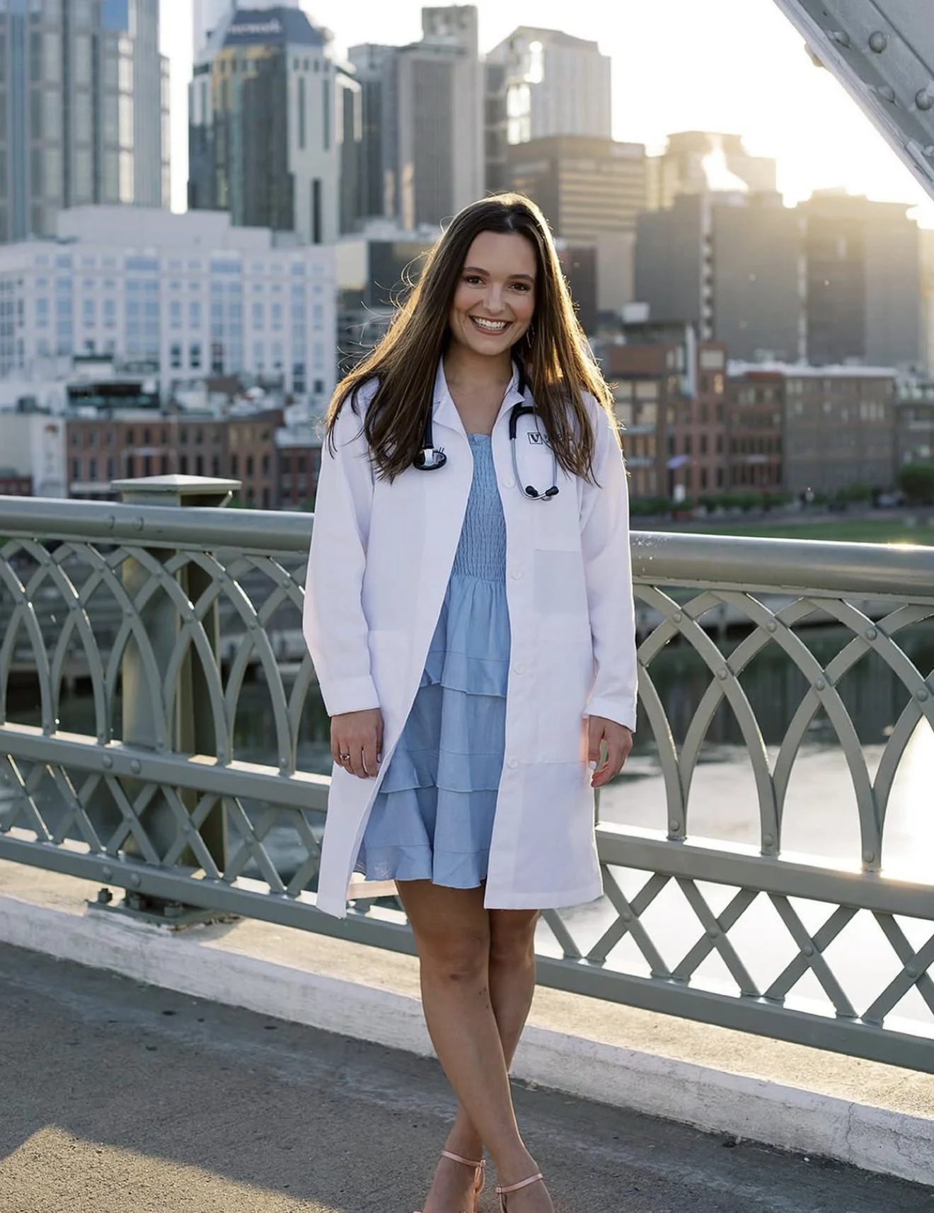A woman in a white doctor's coat and blue dress smiling on a bridge in a city with tall buildings in the background during sunset.