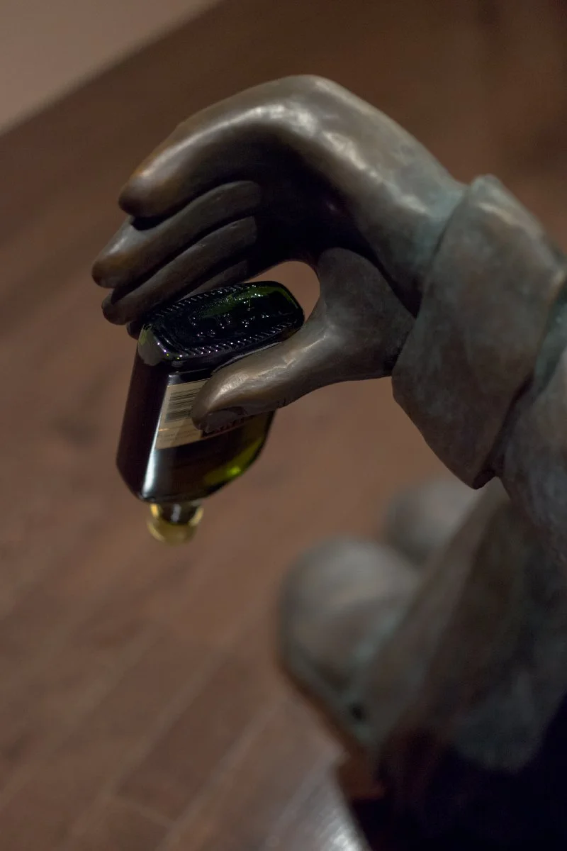 Bronze-colored hand sculpture holding a dark glass bottle upside down.