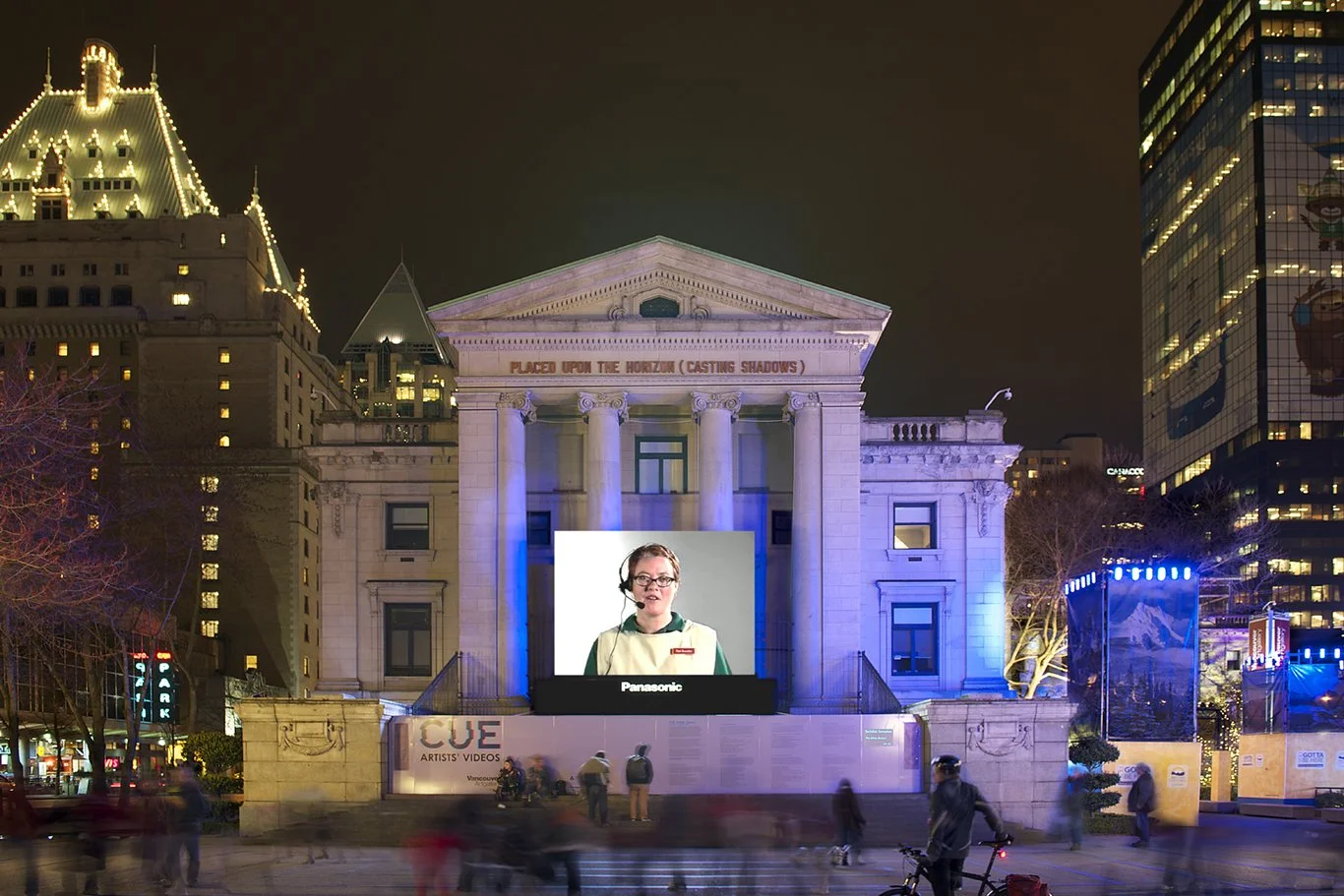 Night scene of a lit historic building with a large digital screen showing a woman in a headset. Surrounding modern high-rise buildings and people walking in the square.