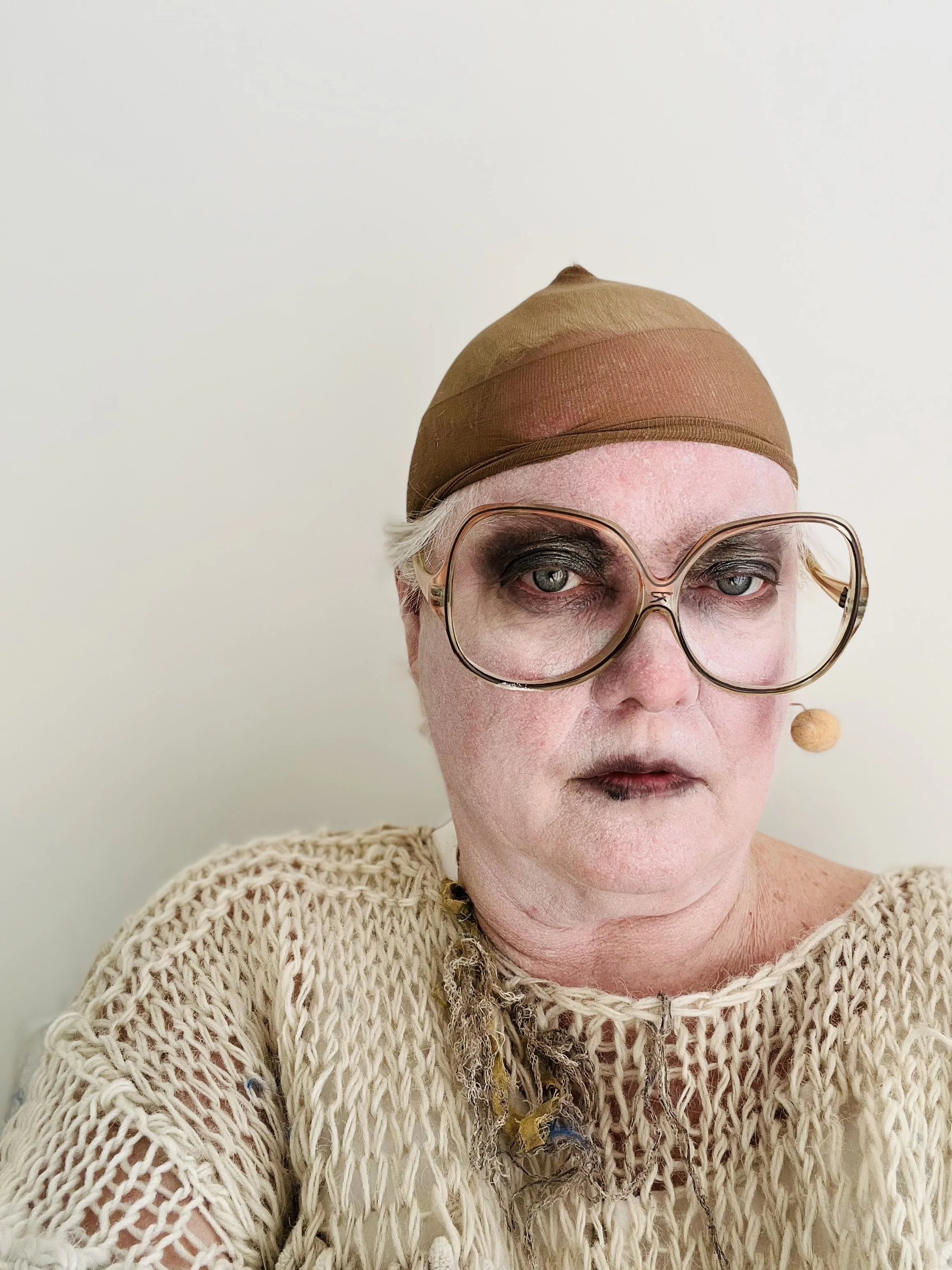 Close-up portrait of a person with white makeup, wearing large glasses, a beige hat, and a cream-colored knit sweater, set against a plain white background.