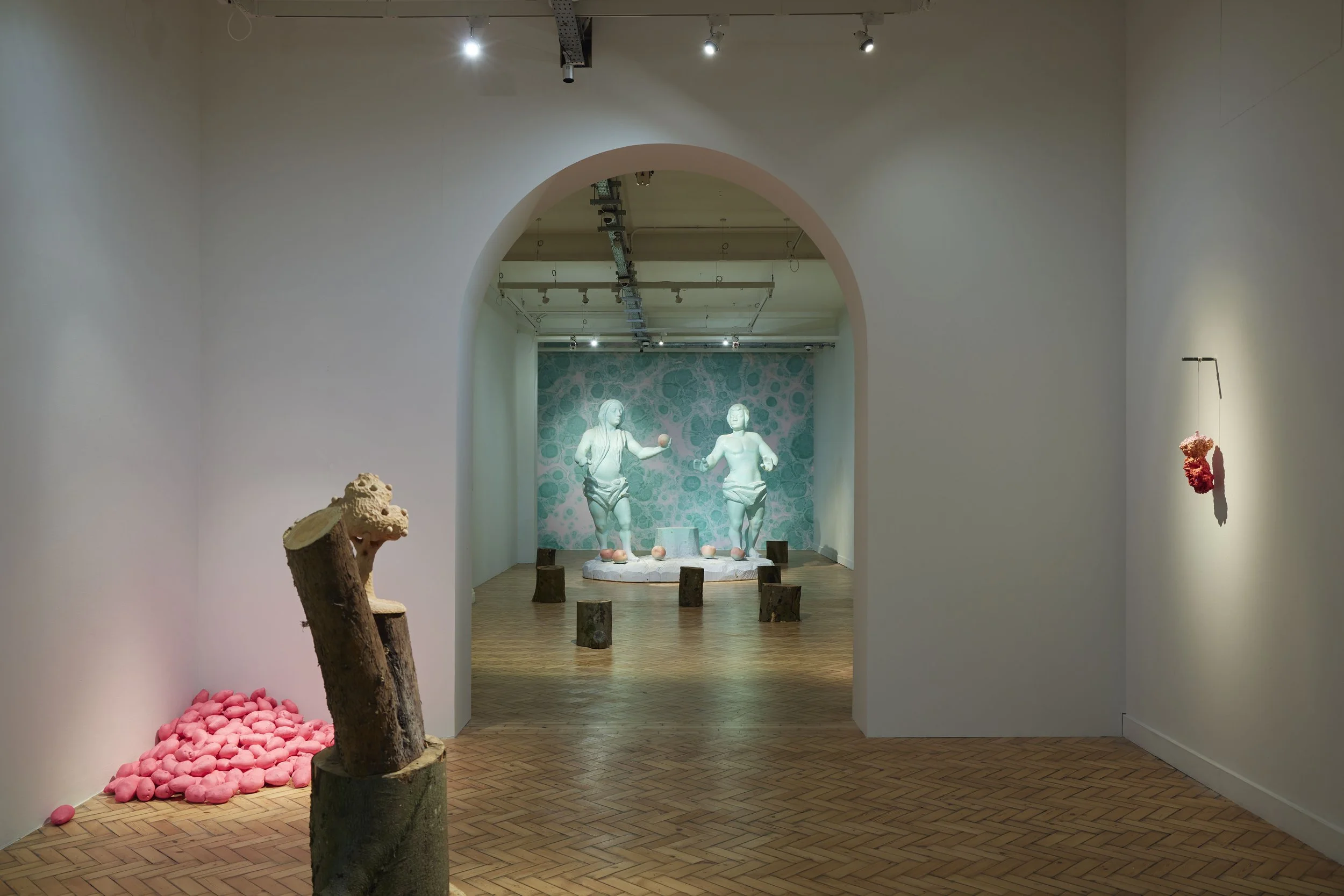 An art installation in a gallery featuring a beige sculpture of Adam and Eve, with a blue marble background. In the foreground, there is a tree stump-like object and pink potatoes on the floor, with ceiling lights illuminating the display.