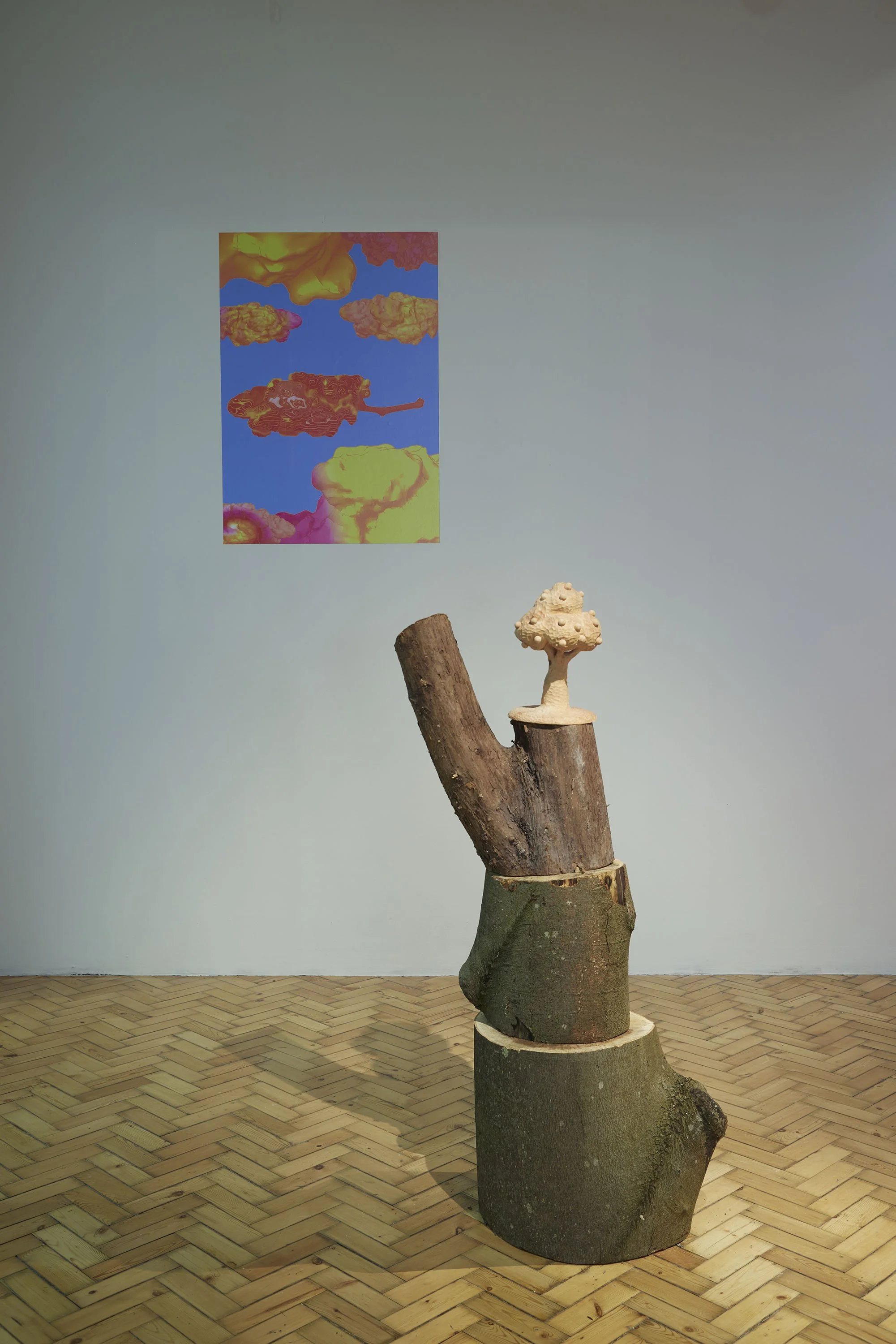 A wooden sculpture of a tree trunk with a small carved wooden tree, placed on a wooden floor with a herringbone pattern. A colourful painting of marijuana buds floating in a blue sky is on the wall behind it.