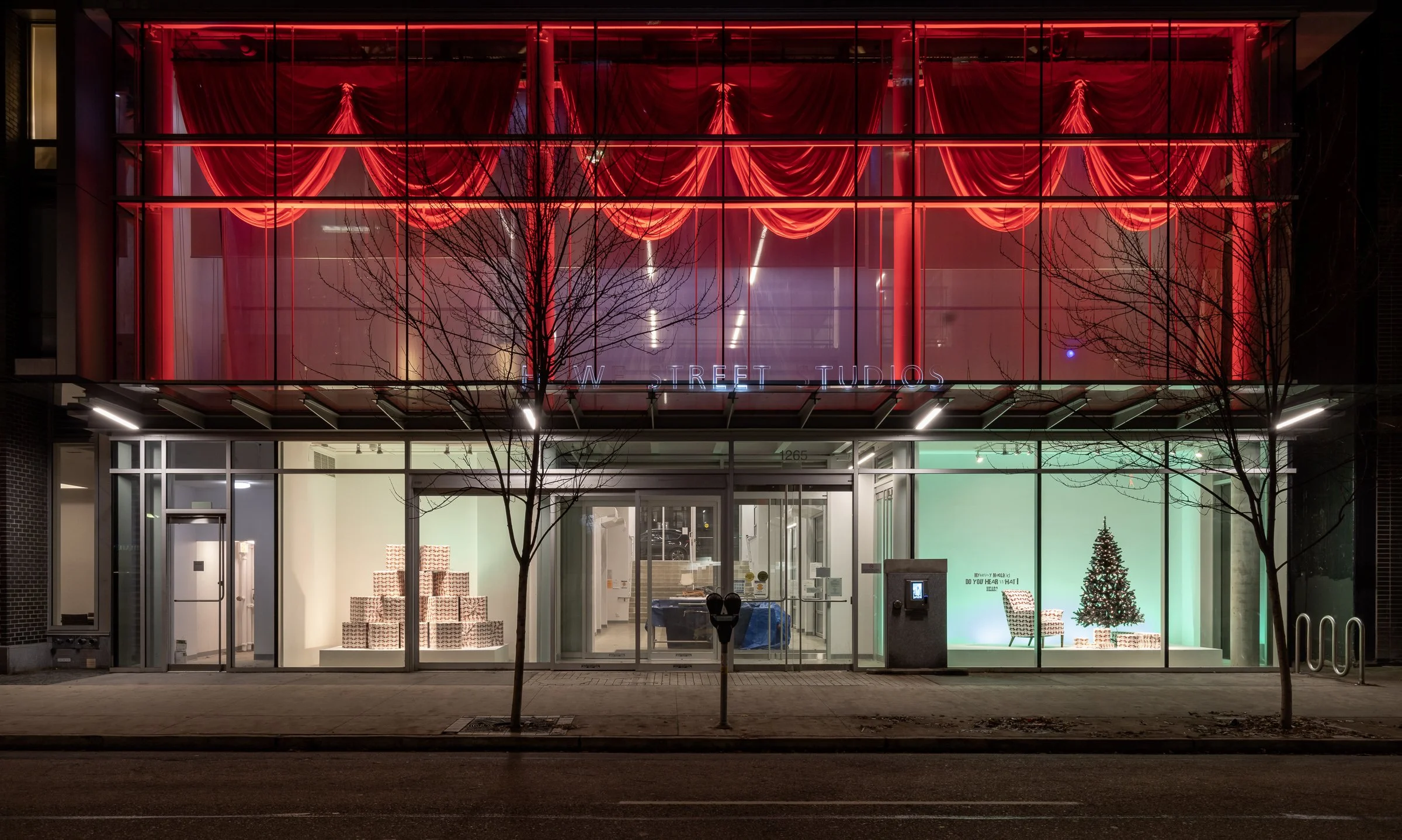 The storefront of a building at night illuminated with bright red neon lights and red curtains behind glass. Inside, there are stacked gift boxes and a small decorated Christmas tree. Outside, there are leafless trees and a sidewalk.