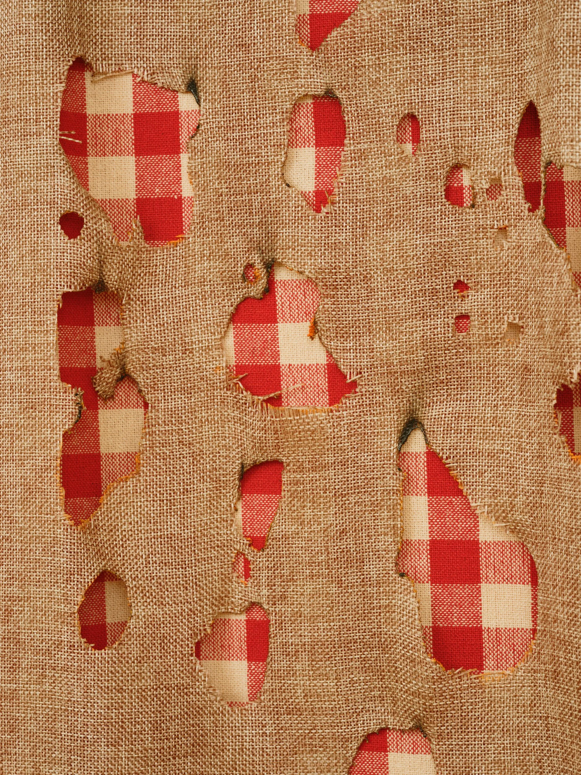 A piece of burlap fabric with several irregular holes, revealing a red and white checkered cloth underneath.