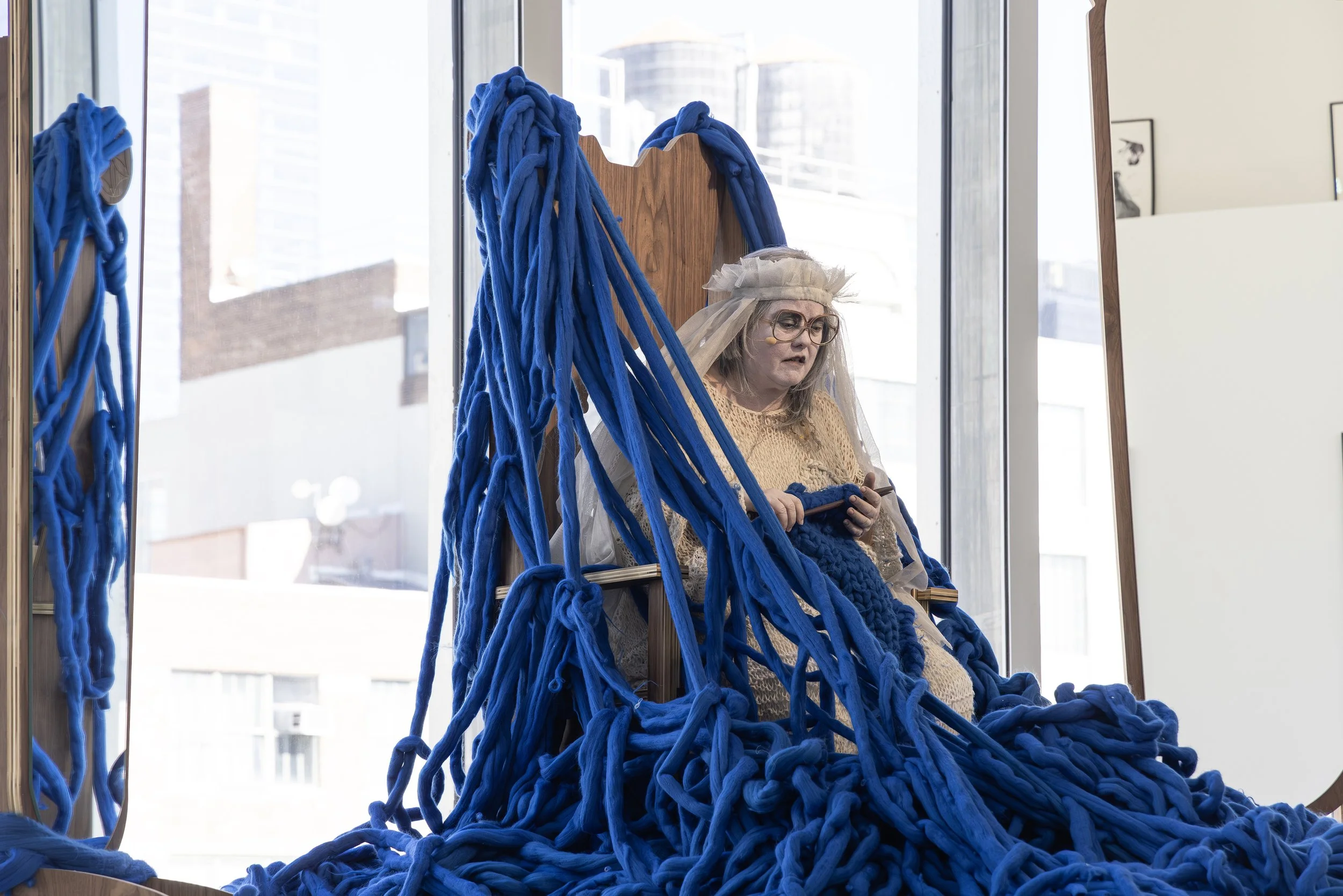 A woman with glasses and a white veil sitting on a chair, surrounded by large blue yarn strands hanging and piled around her, in a room with large windows and city buildings outside.