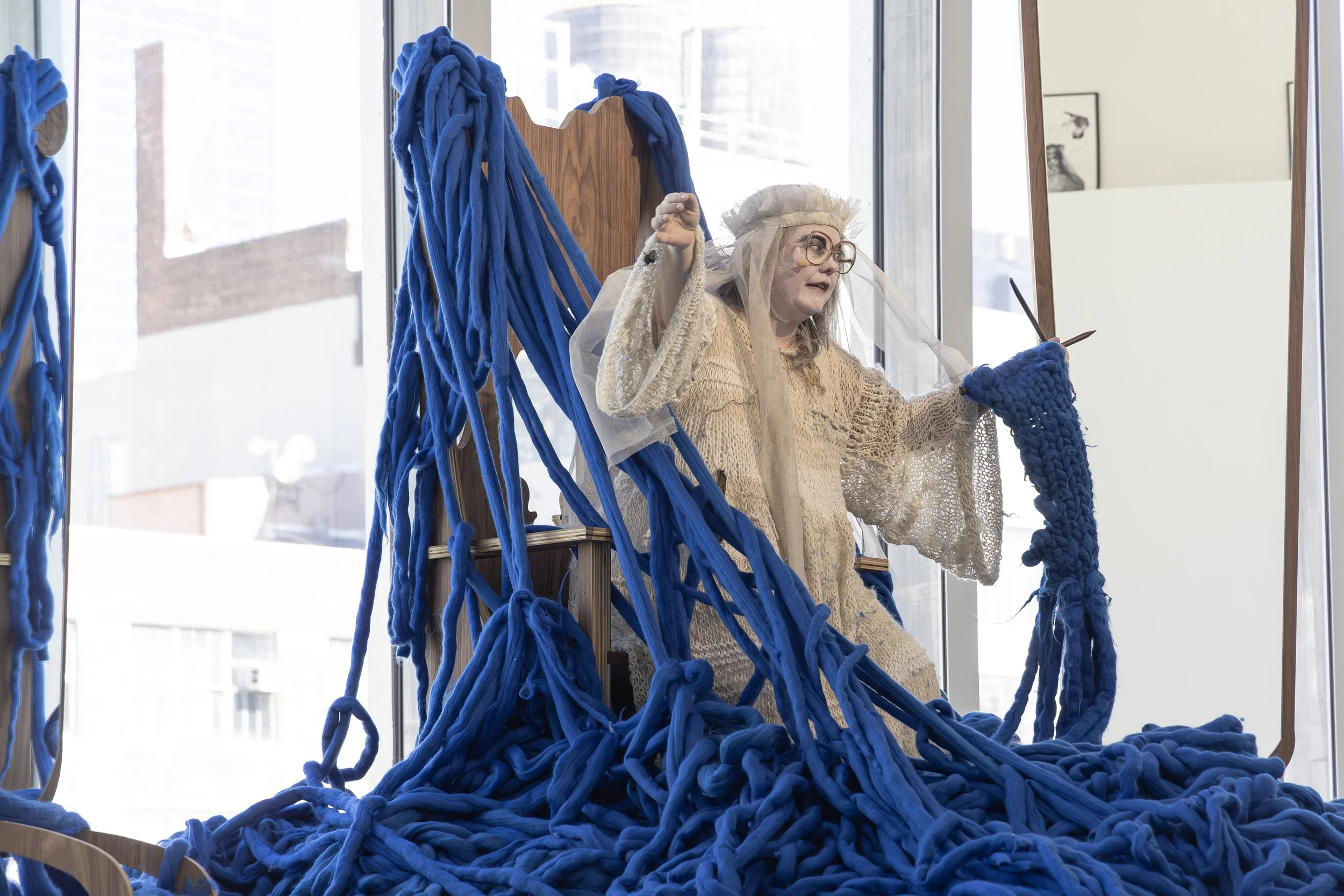 An art installation featuring a person dressed as a woman with gray hair, glasses, and a beige lace dress, surrounded by blue yarn and woven fabric, inside a bright room with large windows and city buildings outside.